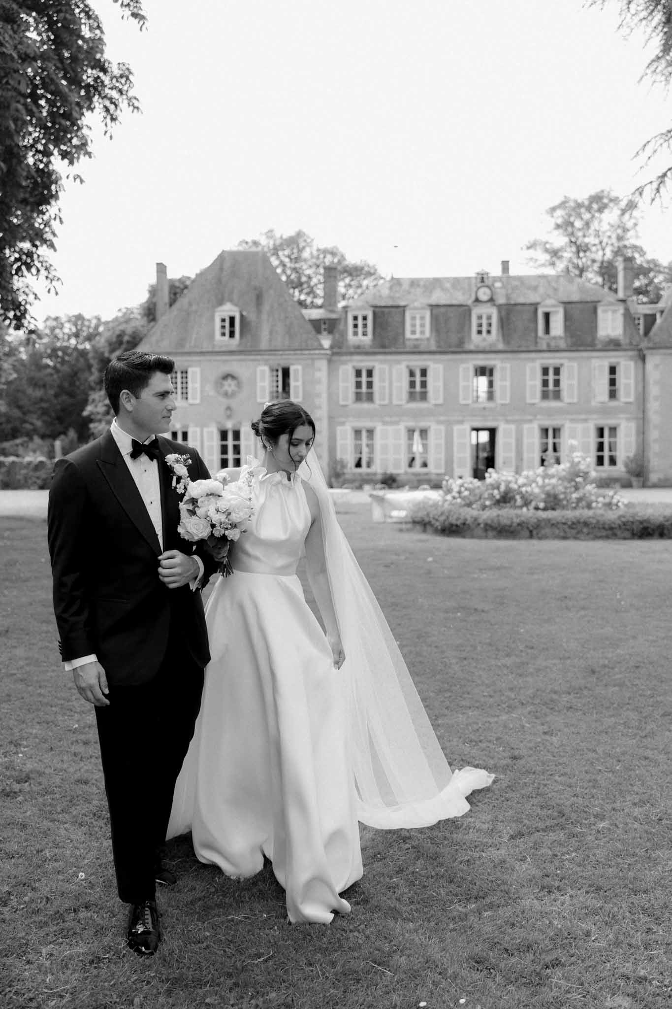 Black-and-white portrait of bride in long-sleeve gown with full skirt and groom on lawn before French château