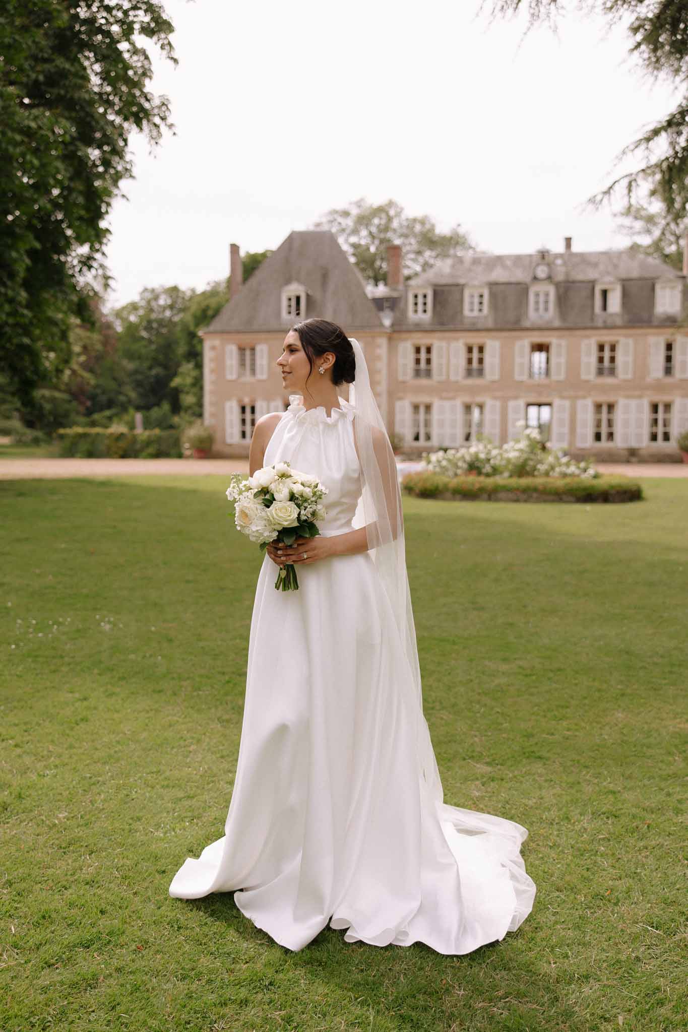 Bride in ivory A-line gown with rose bouquet posing on lawn before a symmetrical ochre stone chateau