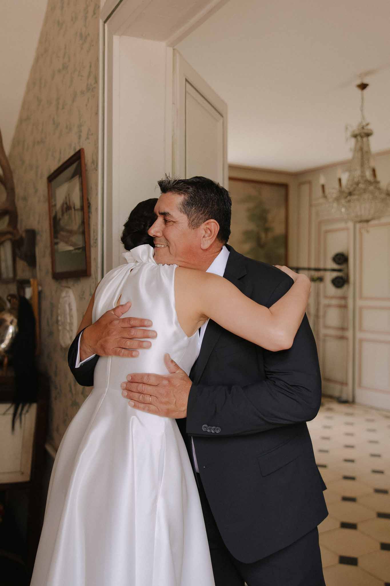 Groom in black suit hugging bride in white sleeveless A-line gown in an elegant hallway with chandelier and classical molding.
