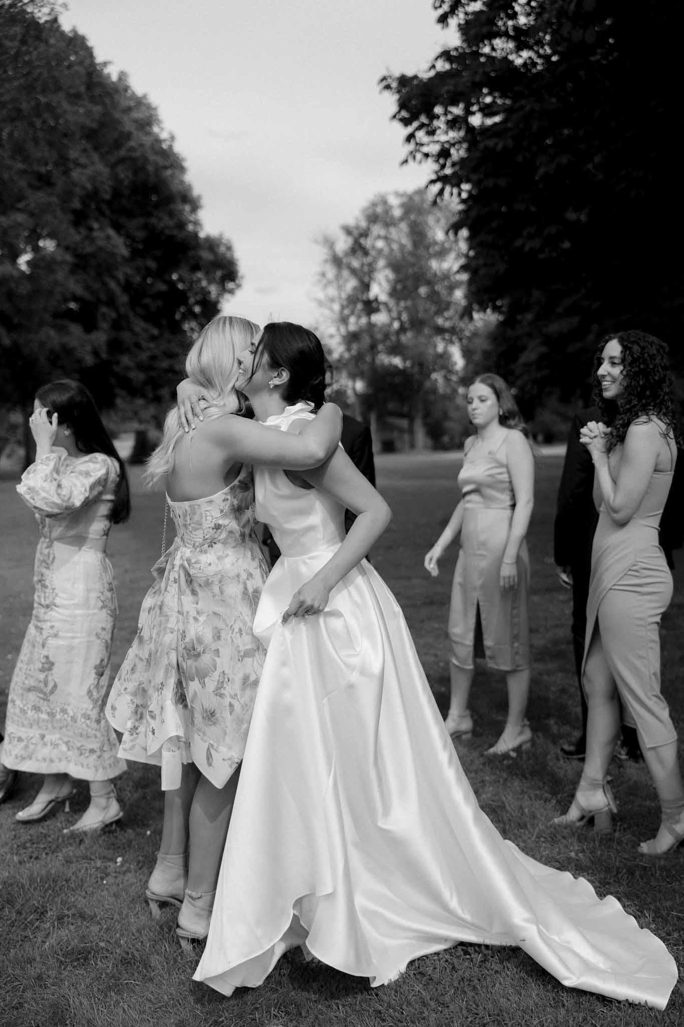 Black and white photo of bride in full skirt embracing a guest surrounded by others in a garden corridor