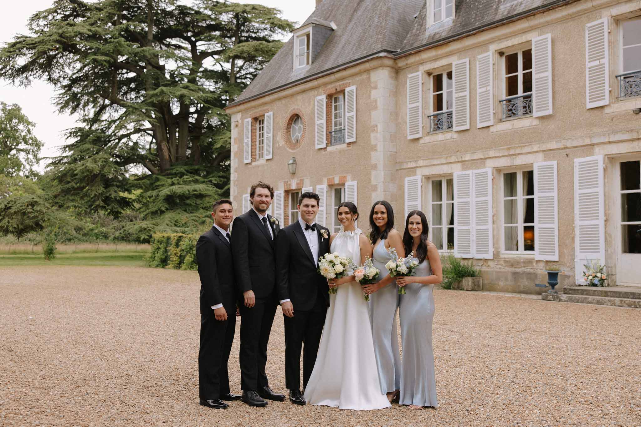 Three groomsmen in black tuxedos, bride in white gown, three bridesmaids in pale gray dresses on gravel courtyard