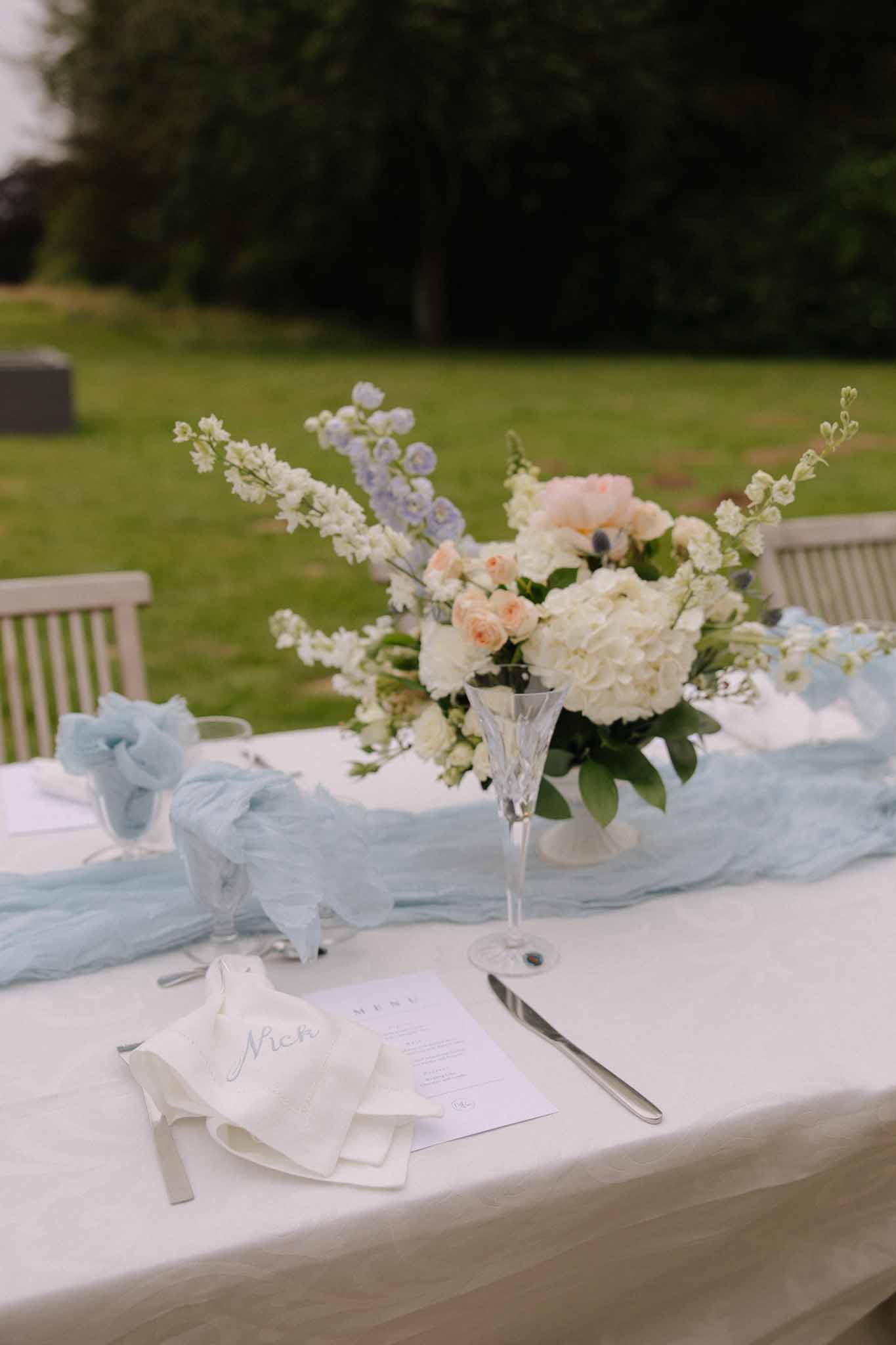 Reception table centerpiece with ivory hydrangeas and peach roses in tall glass vase on blue tulle runner