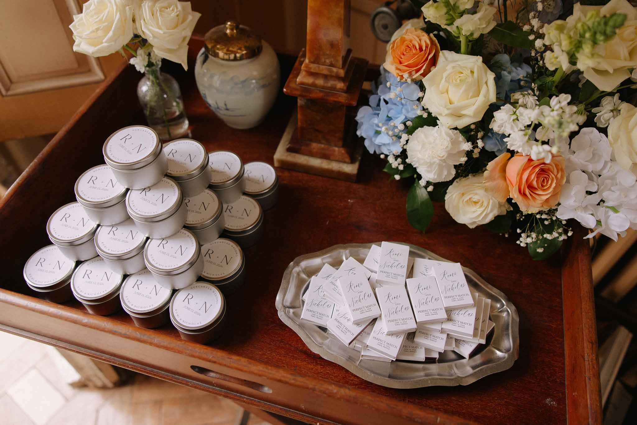 Wedding favours and floral arrangement on dark wood table, white monogrammed tins, escort cards and mixed bouquet