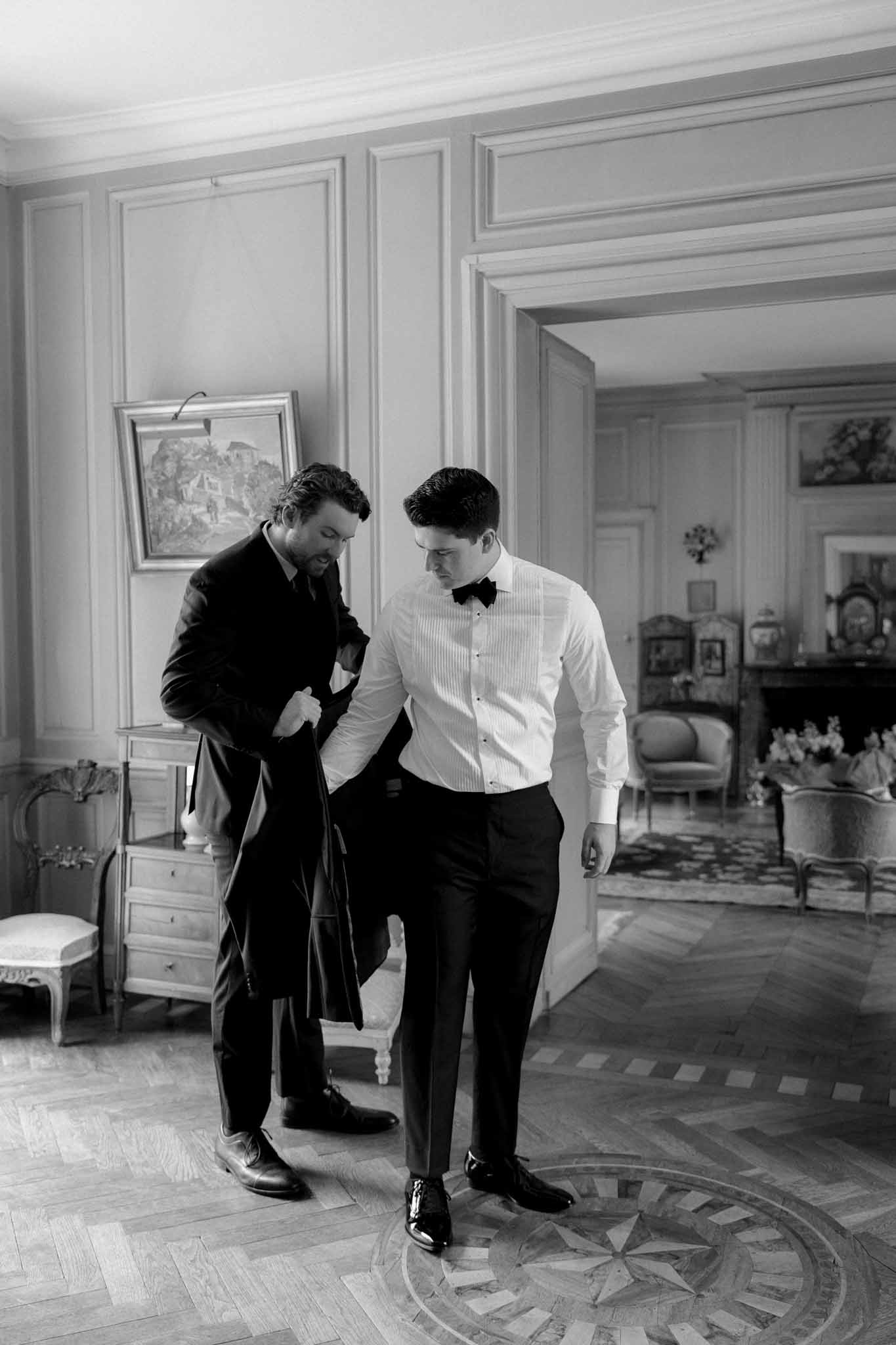 Black and white photo of best man adjusting groom's bow tie in classical room with parquet floor