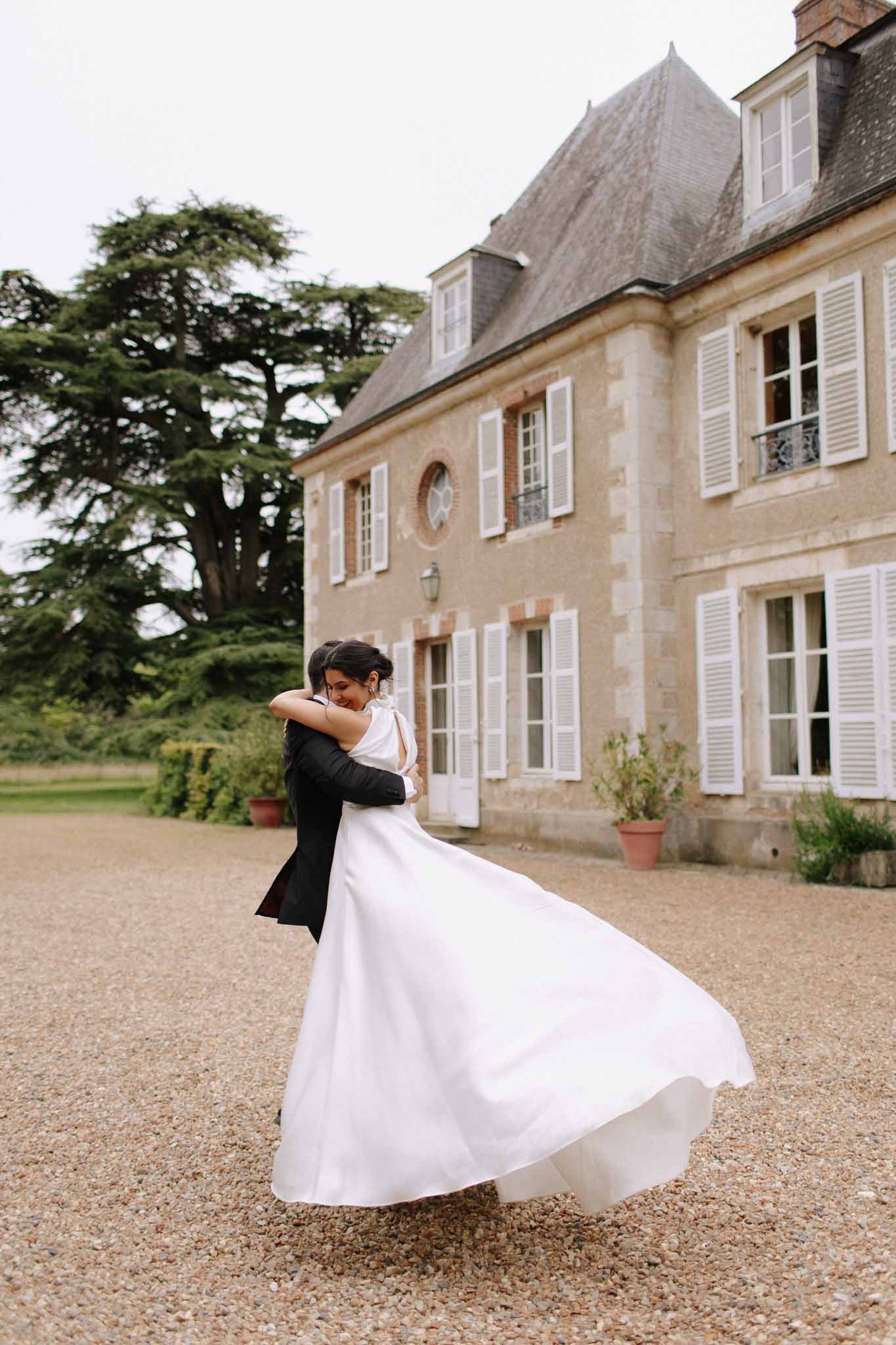 Groom spinning bride on gravel courtyard in front of classical French stone manor with mansard roof