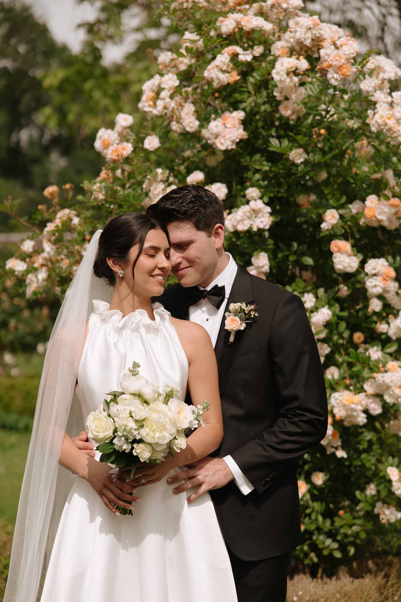 Bride and groom embracing in front of climbing peach and ivory roses on garden wall