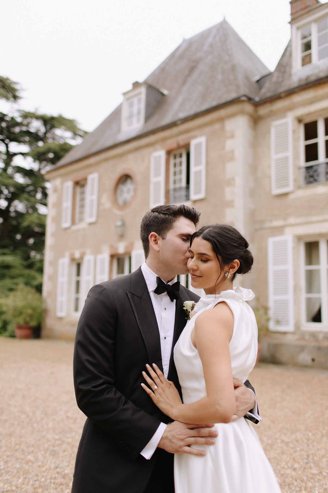 Bride and groom in courtyard of stone château, groom in black tuxedo, bride in white A-line gown with bow detail