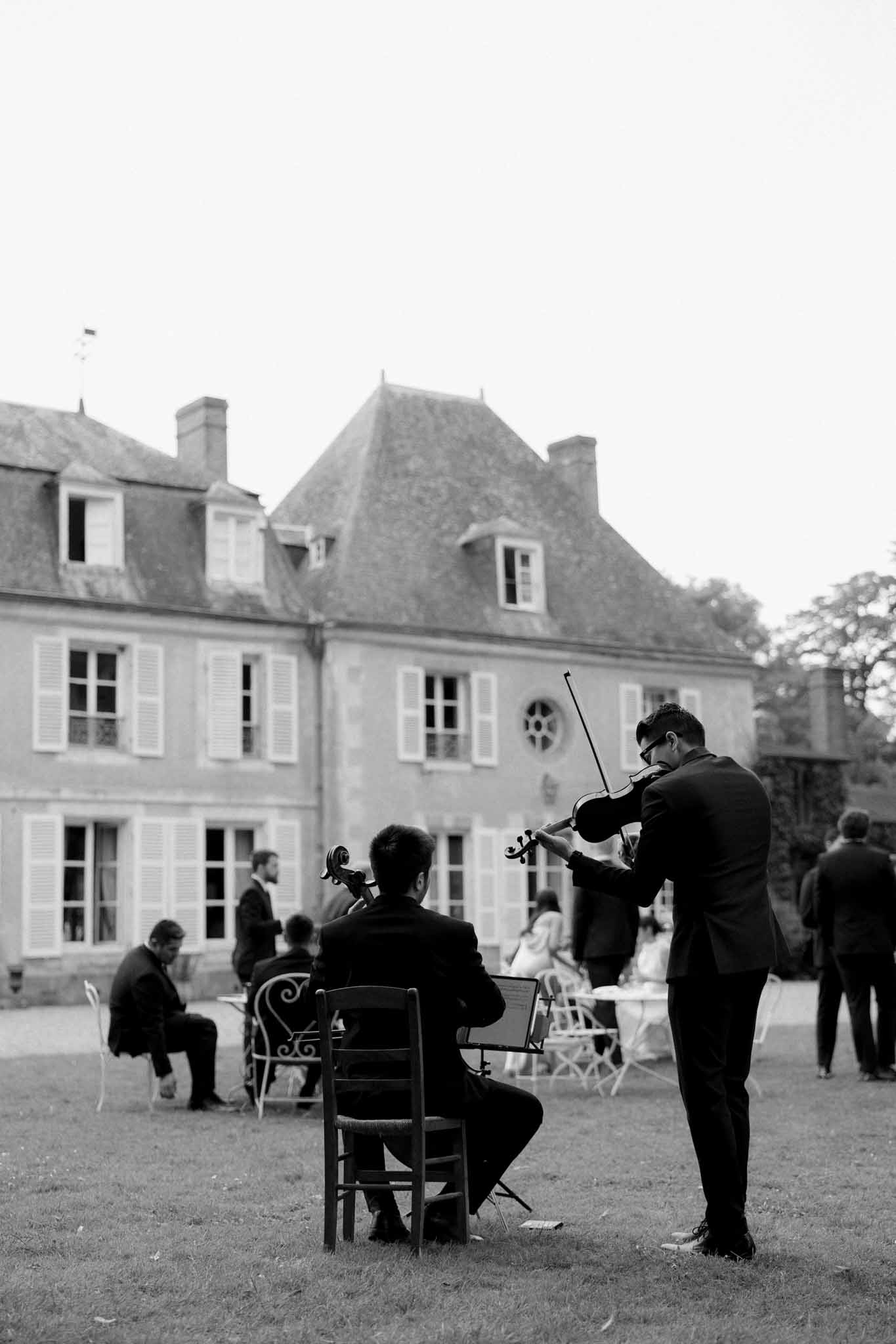 Black and white photo of two musicians playing violin and cello for guests in the courtyard of a French château
