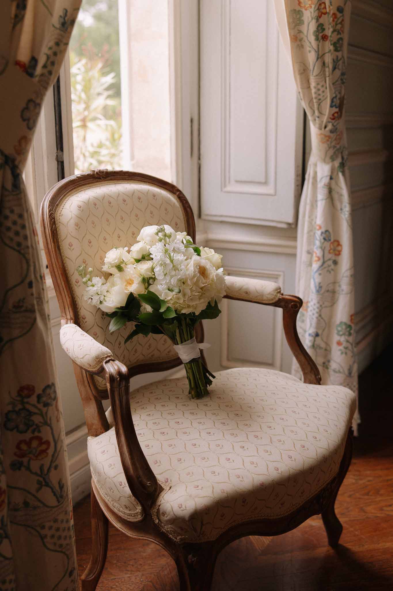 Ivory peony and white spray rose bridal bouquet resting on carved Louis XVI-style chair, botanical print curtains behind