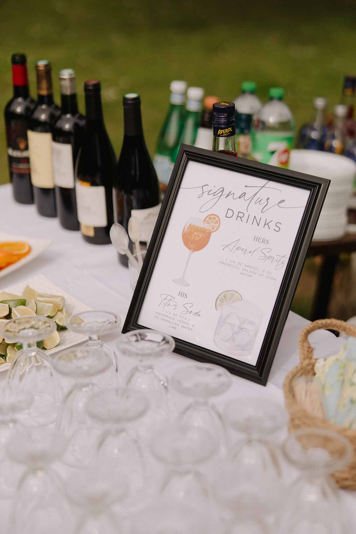 Signature drinks sign with handwritten cocktail recipes beside coupe glasses and Aperol bottle on white tablecloth