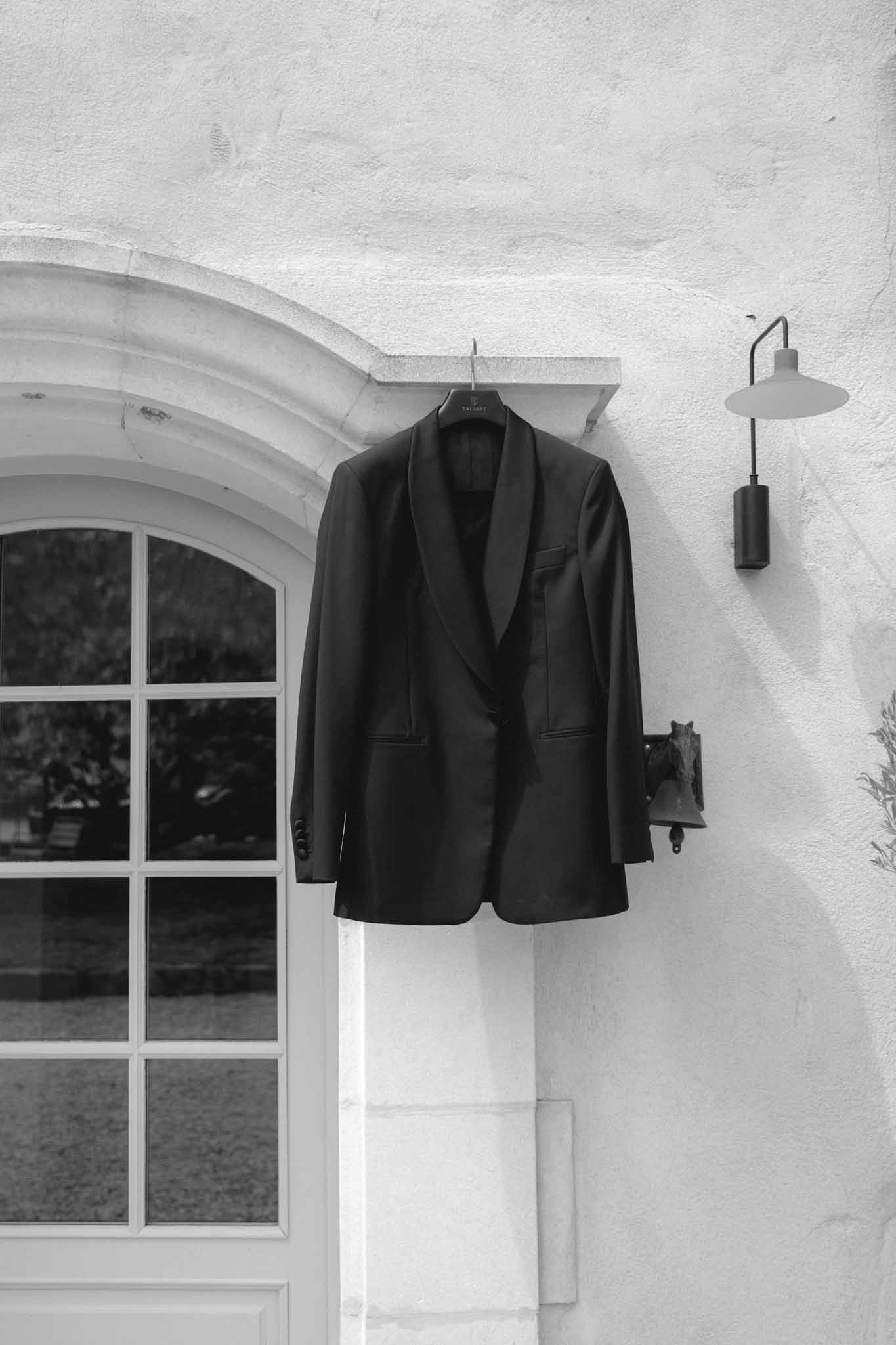 Black blazer hanging on metal hanger above arched window on whitewashed exterior wall, black-and-white photograph