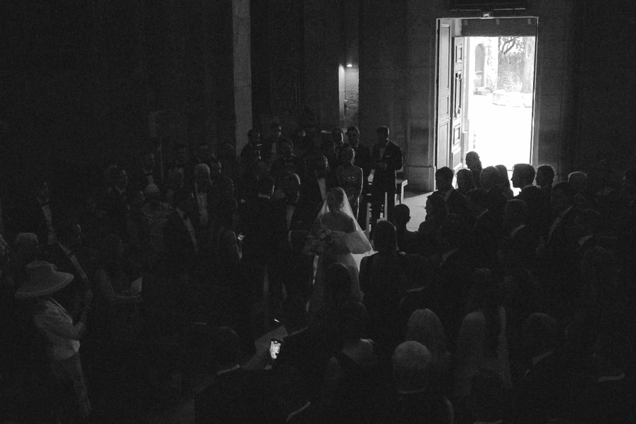 Black and white photo of bride's entrance in a stone chapel corridor with guests standing on both sides