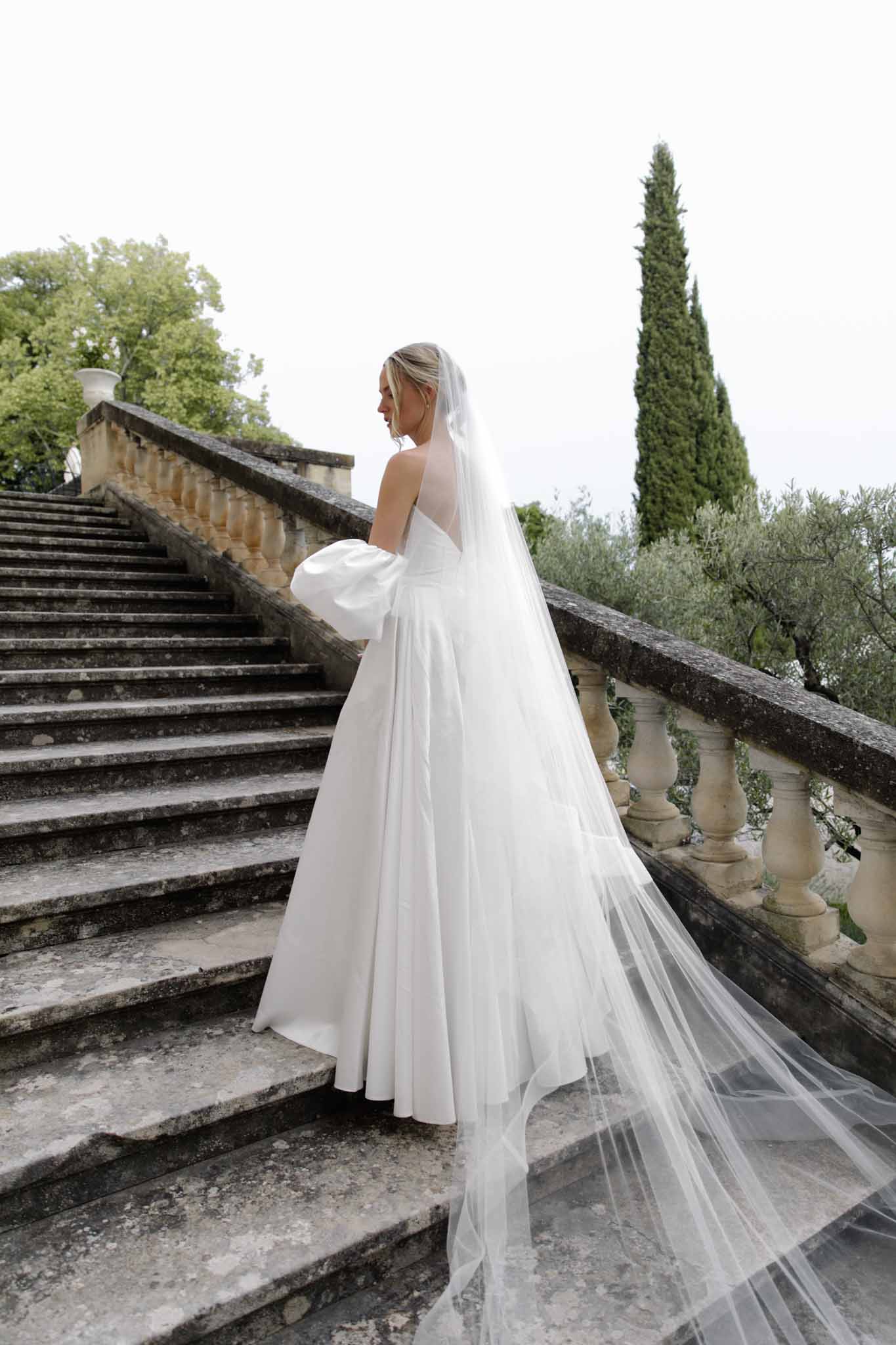 Bride in ivory A-line gown in profile on a stone staircase with long veil trailing down the steps; classical stone balustrade.