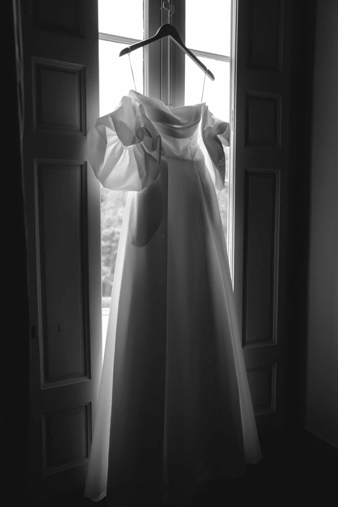 Bride's wedding gown hanging on black hanger in front of tall window with dark wooden doors, black and white