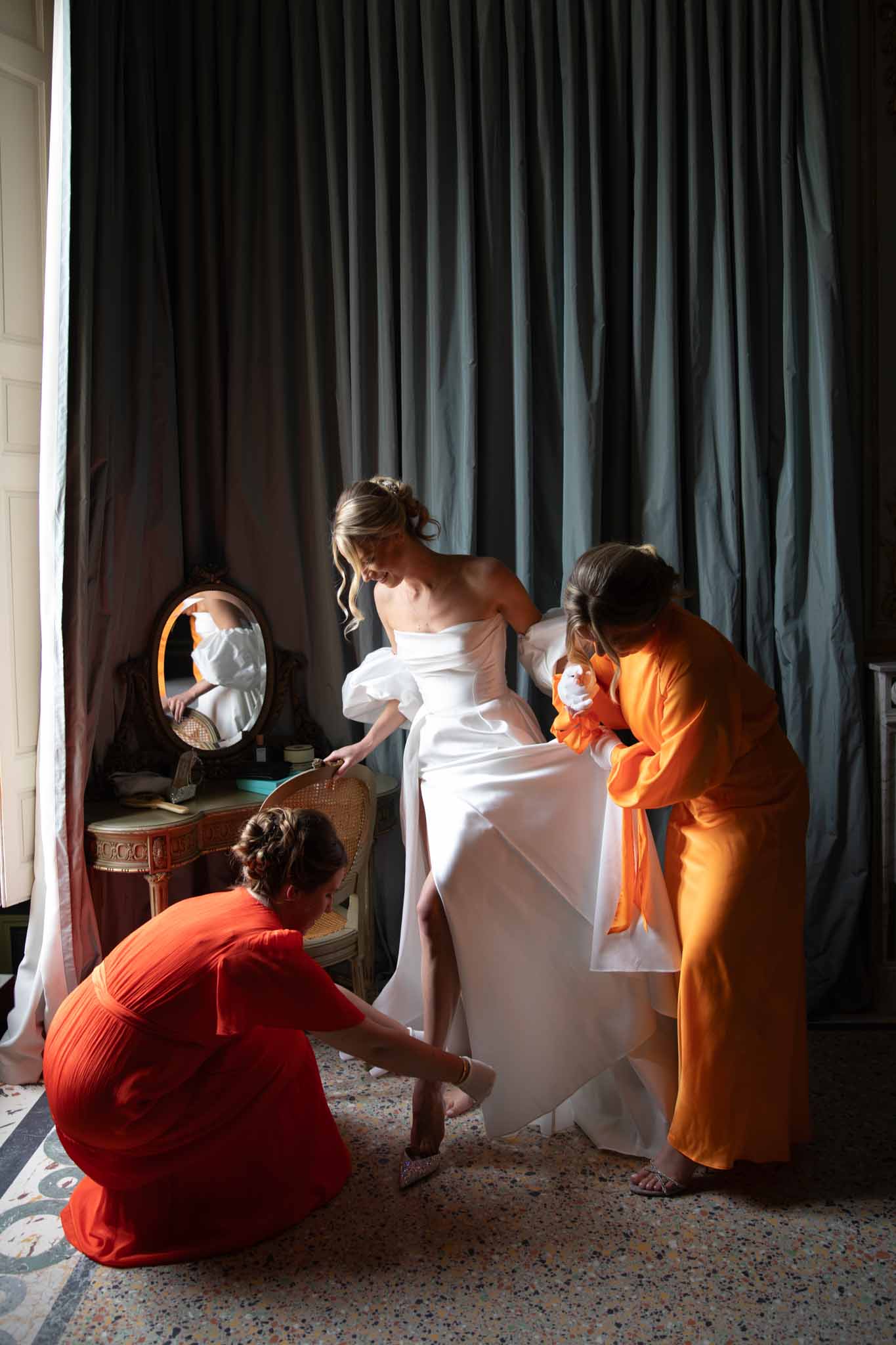 Bride in ivory strapless gown attended by three women in jewel-toned robes adjusting her dress in room with teal velvet curtains