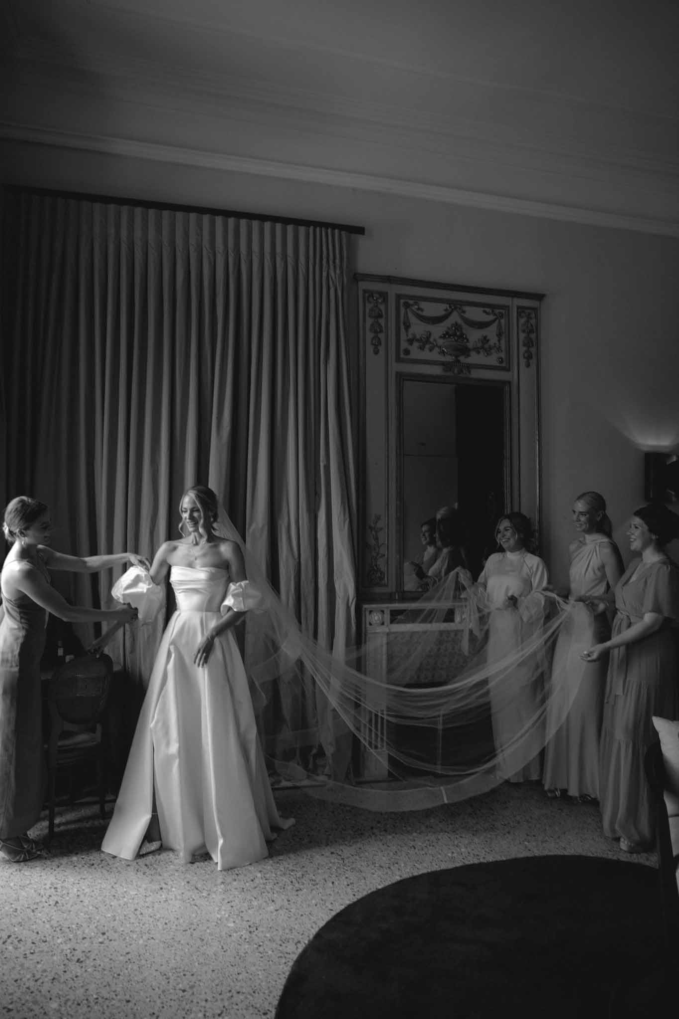 Black-and-white photo of bride with seven bridesmaids arranging veil and train in classical interior room