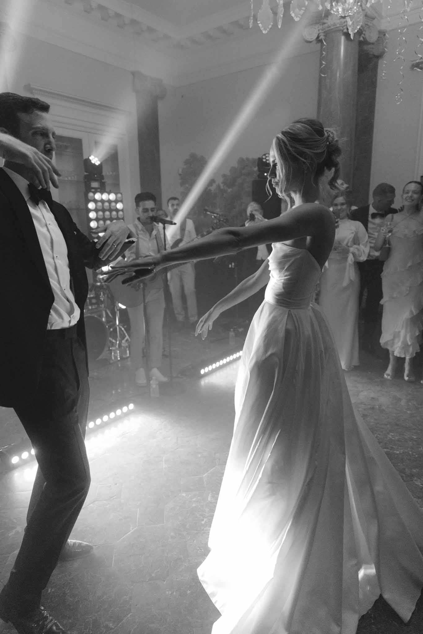 Black and white first dance in a ballroom; bride in strapless gown and groom in tuxedo, live band and guests visible behind.