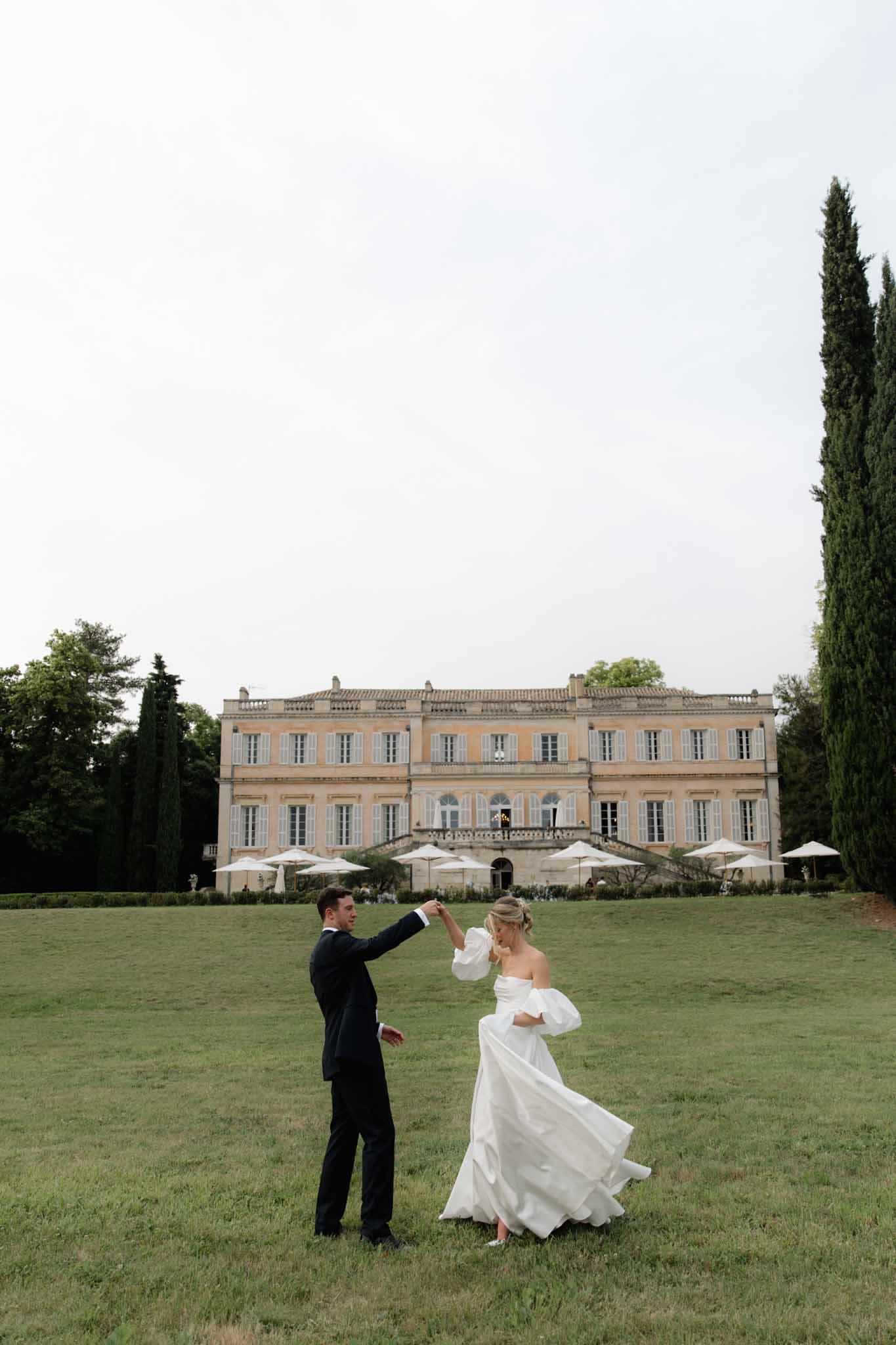 Bride and groom dancing on lawn in front of neoclassical chateau with cypress trees and lounge areas