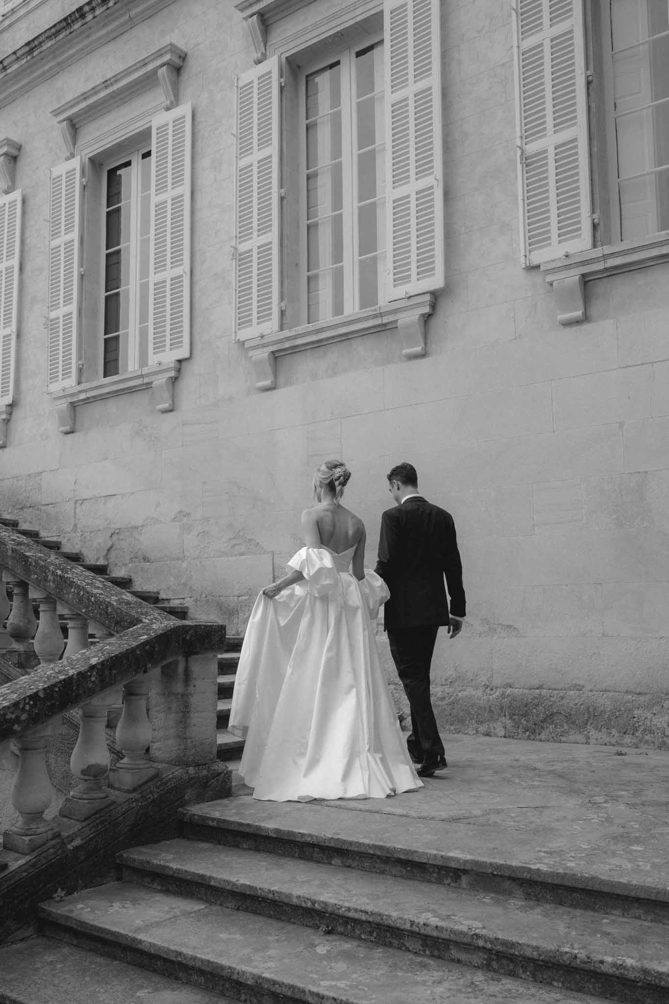 Black and white photo of bride and groom ascending stone steps at a neoclassical chateau courtyard from behind