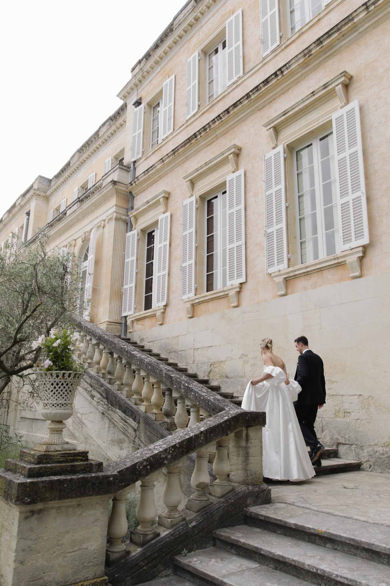 Bride and groom walking up exterior stone staircase of honey-colored 18th-century French chateau with white shutters