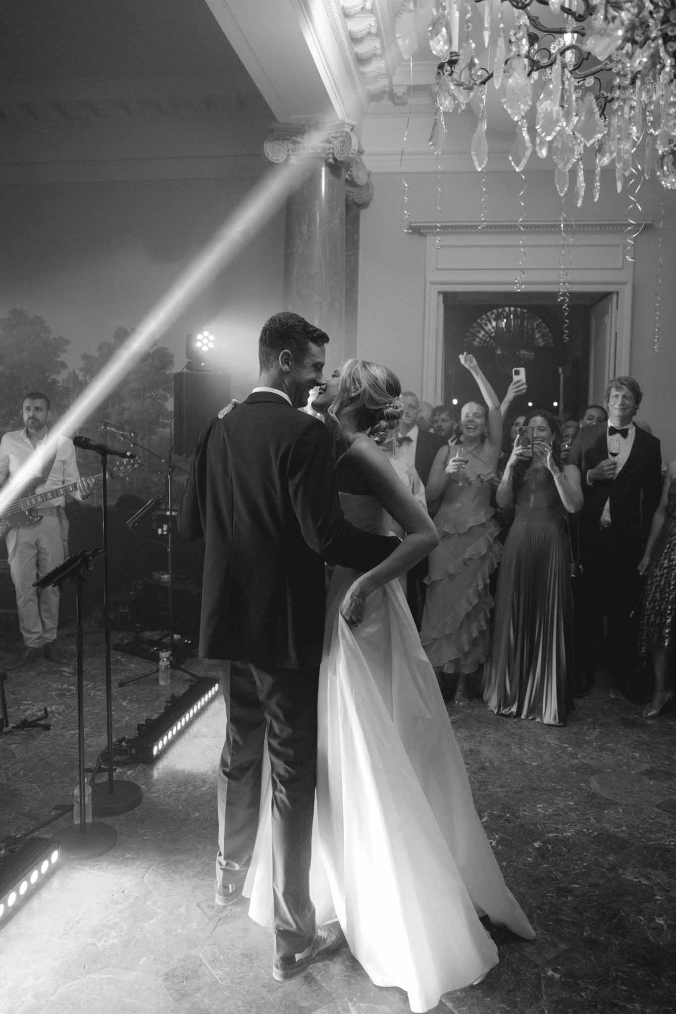 First dance in classical ballroom, black and white, bride in A-line gown, groom in dark suit, live band and guests watching