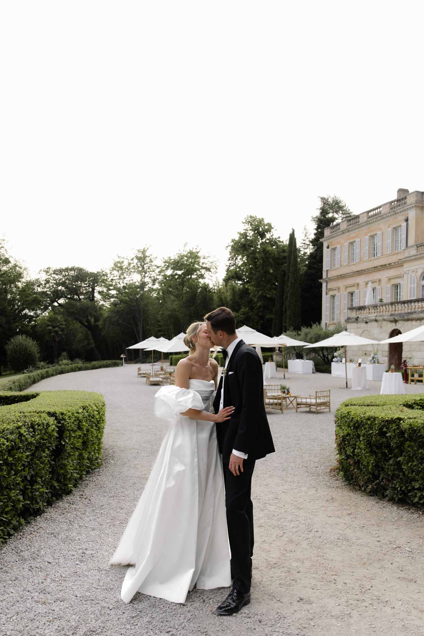 Bride and groom kiss on gravel courtyard pathway at château estate, white tent and gold chairs visible in background