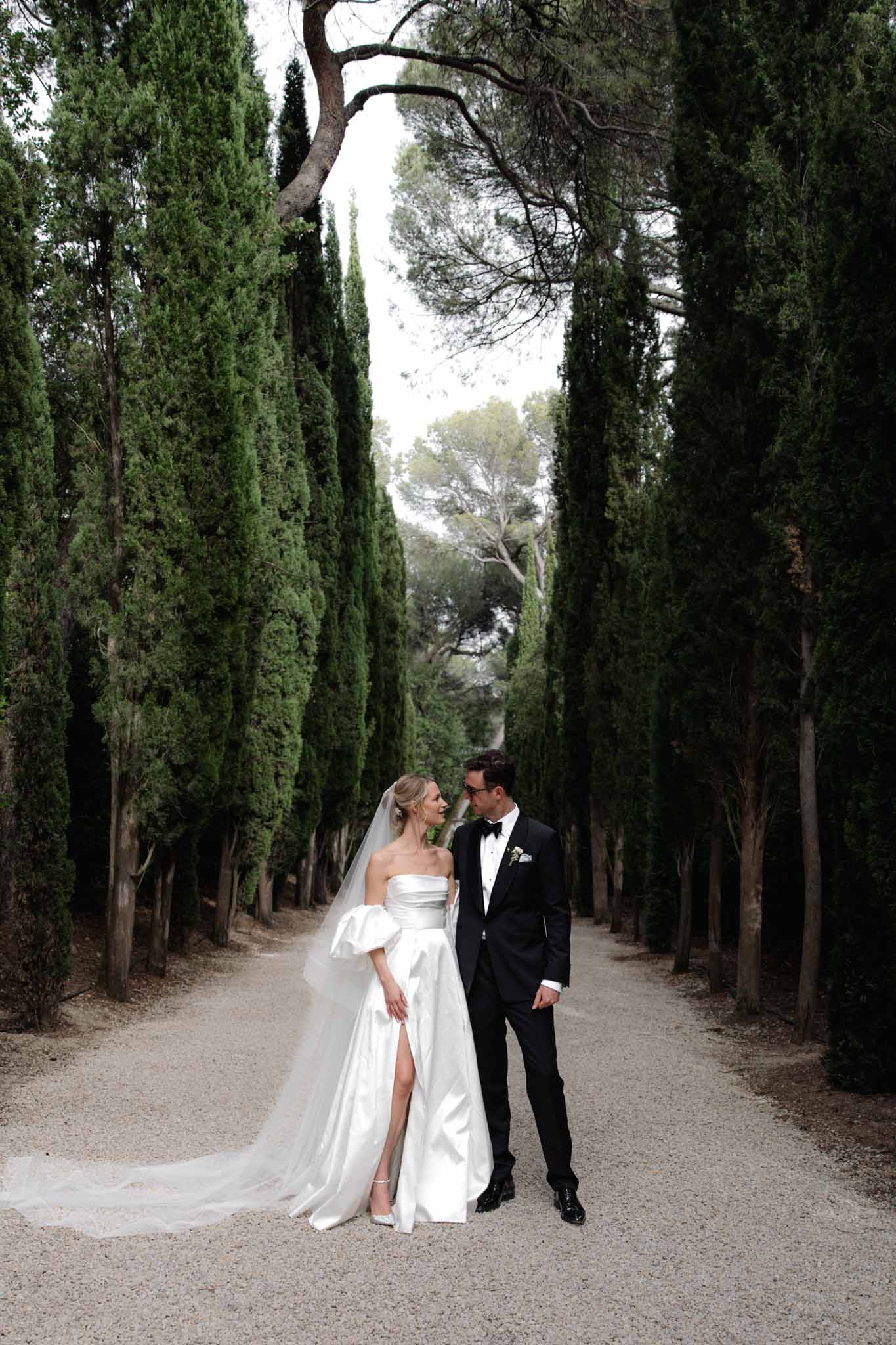 Bride in off-shoulder satin gown with veil and groom in tuxedo walking down cypress tree-lined avenue