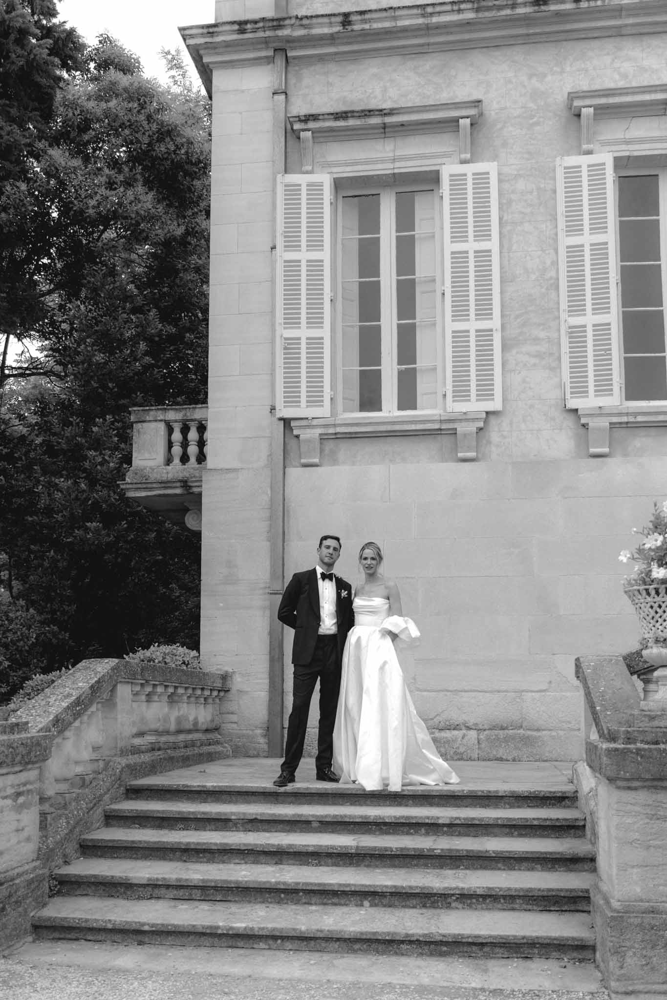 Black-and-white portrait of bride in strapless gown and groom on stone staircase before neoclassical stone mansion