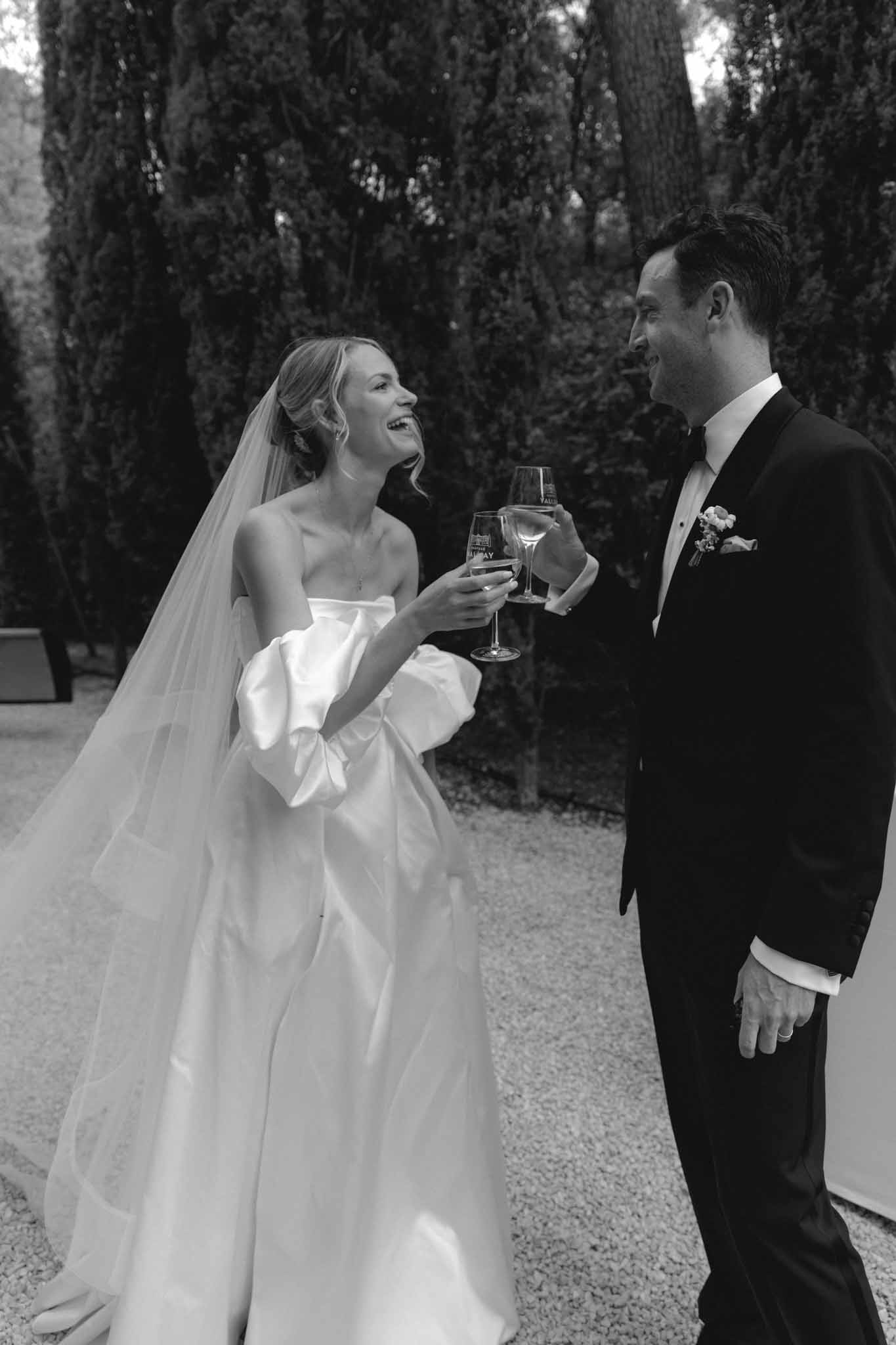 Bride and groom toasting with champagne glasses on gravel path lined with tall cypress trees, black and white photo