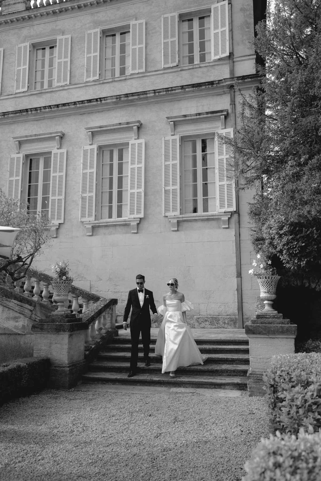 Black-and-white portrait of bride in off-shoulder gown and groom in tuxedo with sunglasses descending château stone steps
