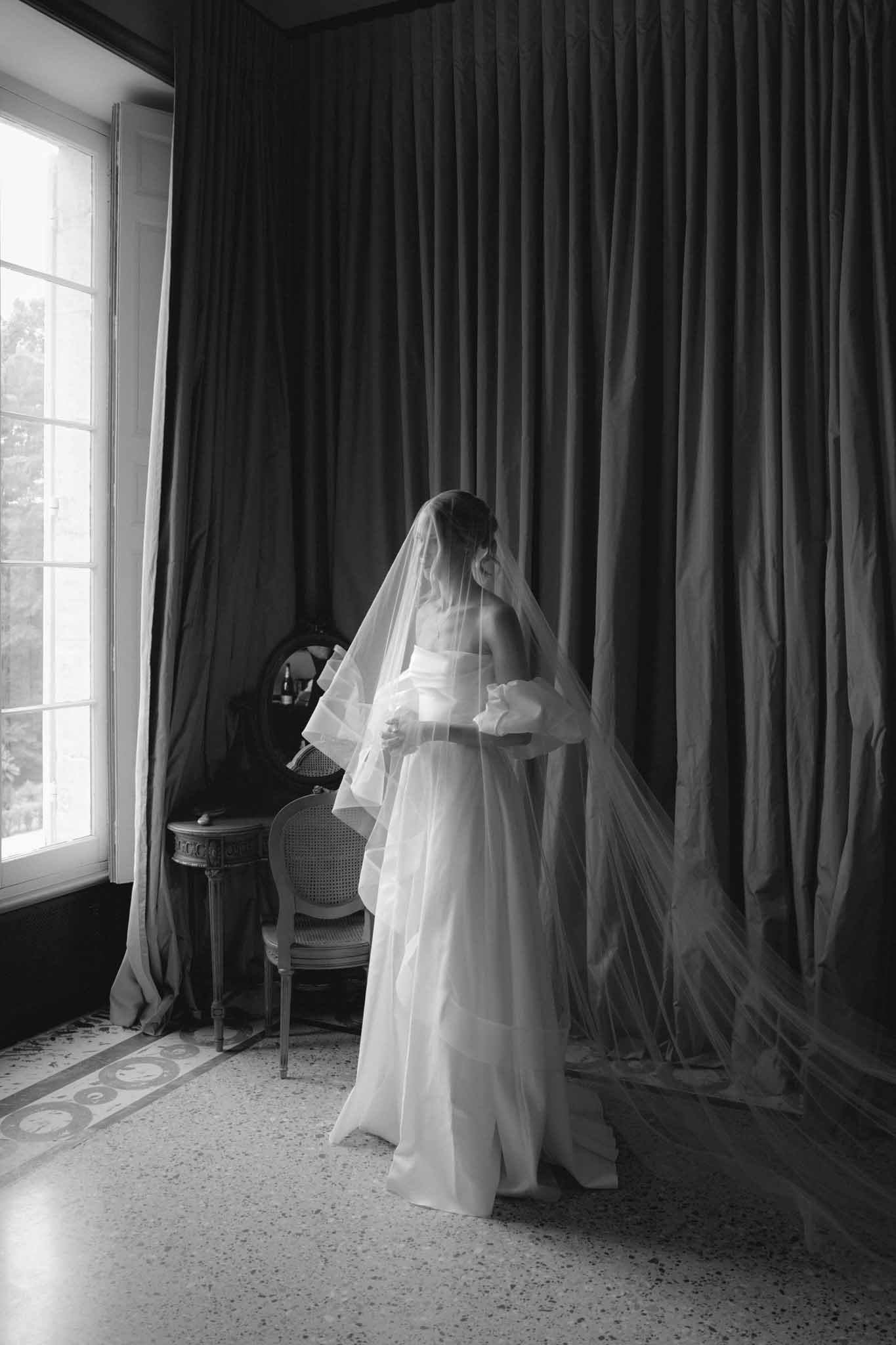 Black and white portrait of bride by window in flowing gown and long veil during getting ready