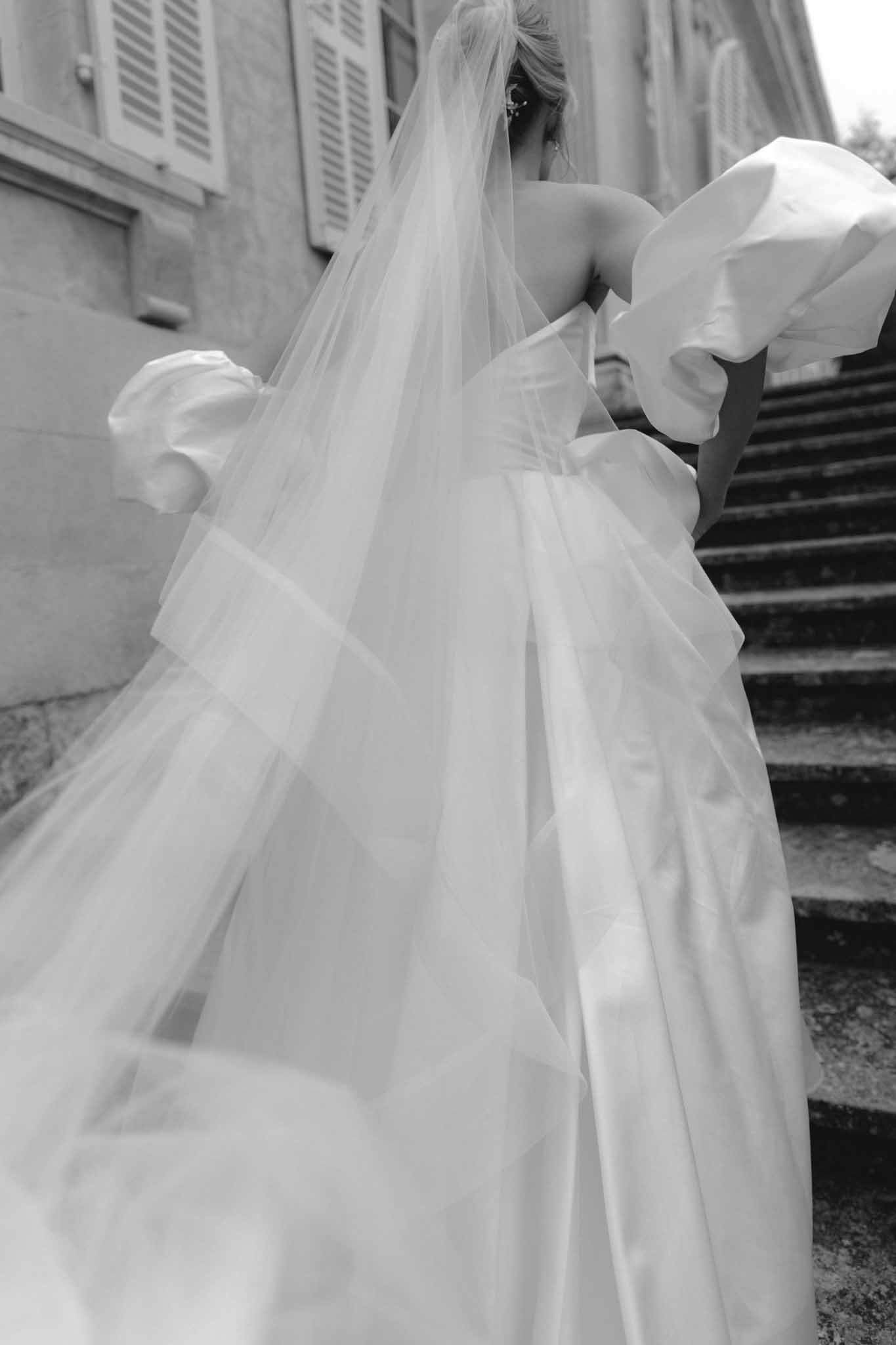 Black and white portrait of bride ascending stone stairs with dramatic tulle veil flowing down the steps