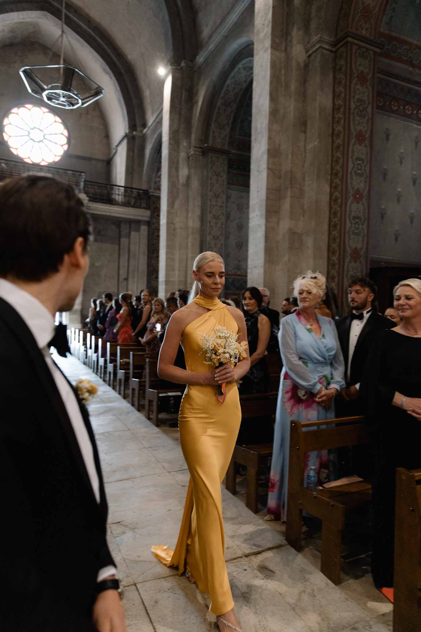 Woman in gold halter gown walks ceremony aisle in historic chapel with stone columns and neoclassical painted walls