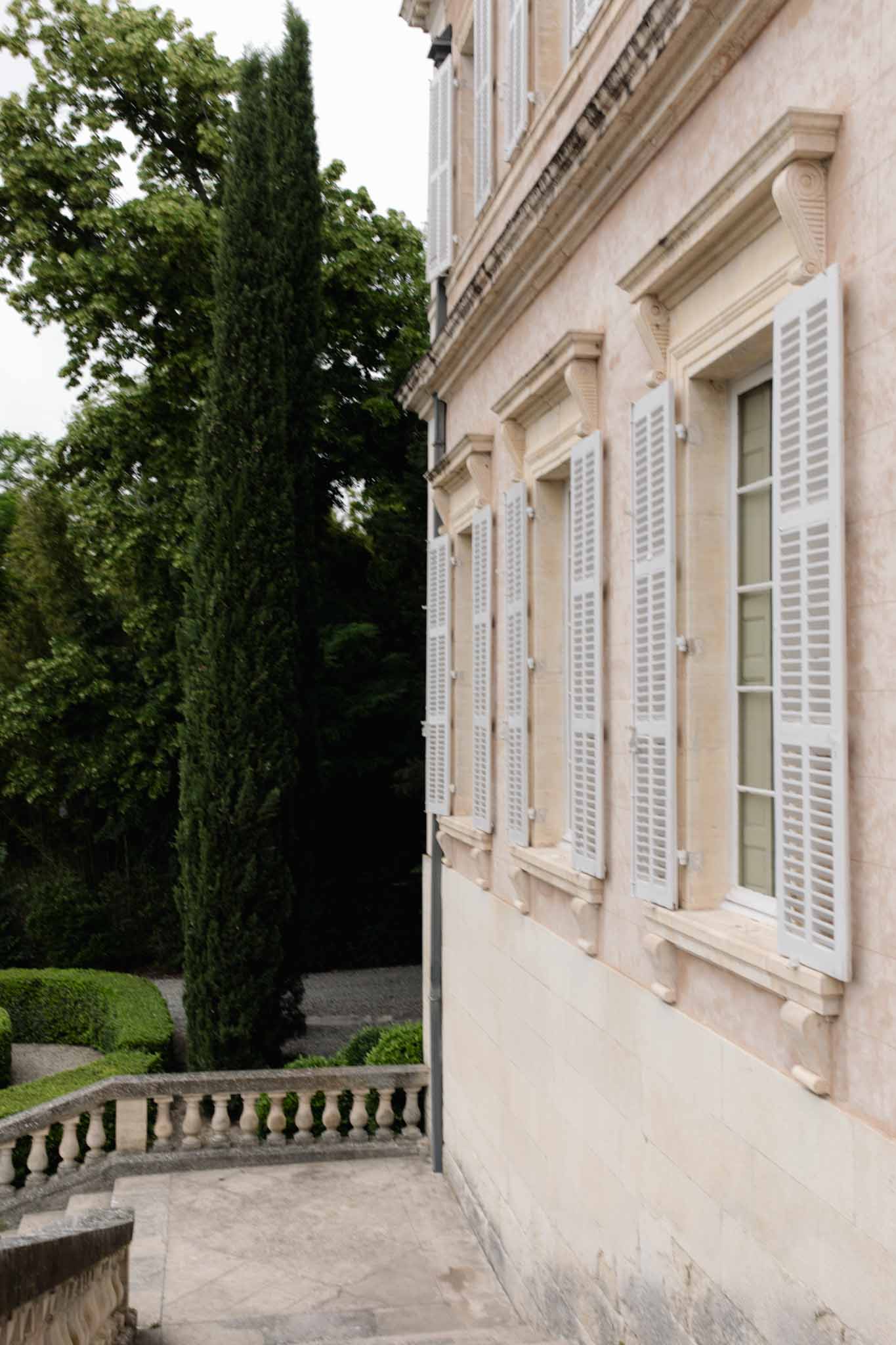 Neoclassical chateau facade with cream walls, white louvered shutters, and cypress trees framing the terrace