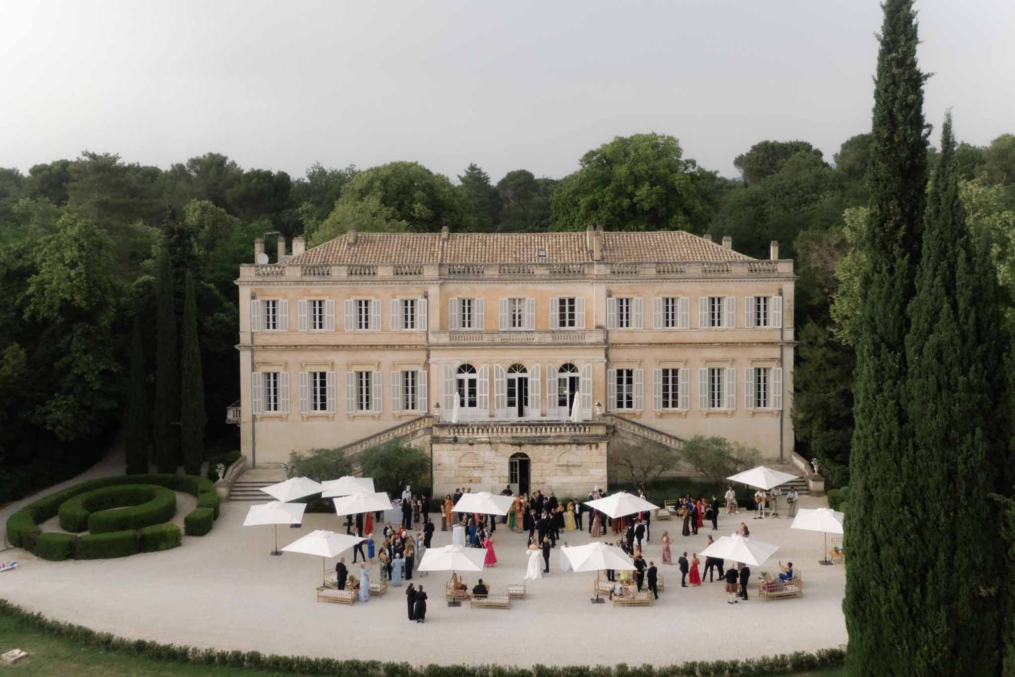 Aerial view of a classical French château courtyard with 150 guests under white umbrellas, cypress trees flanking the facade.