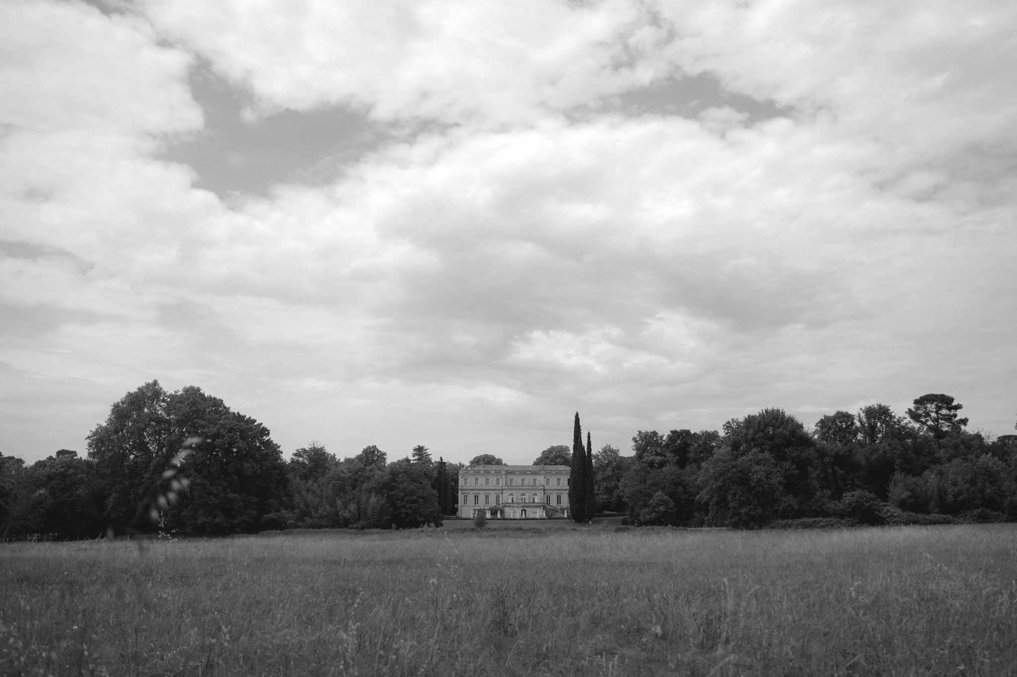 Black and white photograph of Chateau de Martinay across an open field with cypress trees and dramatic sky