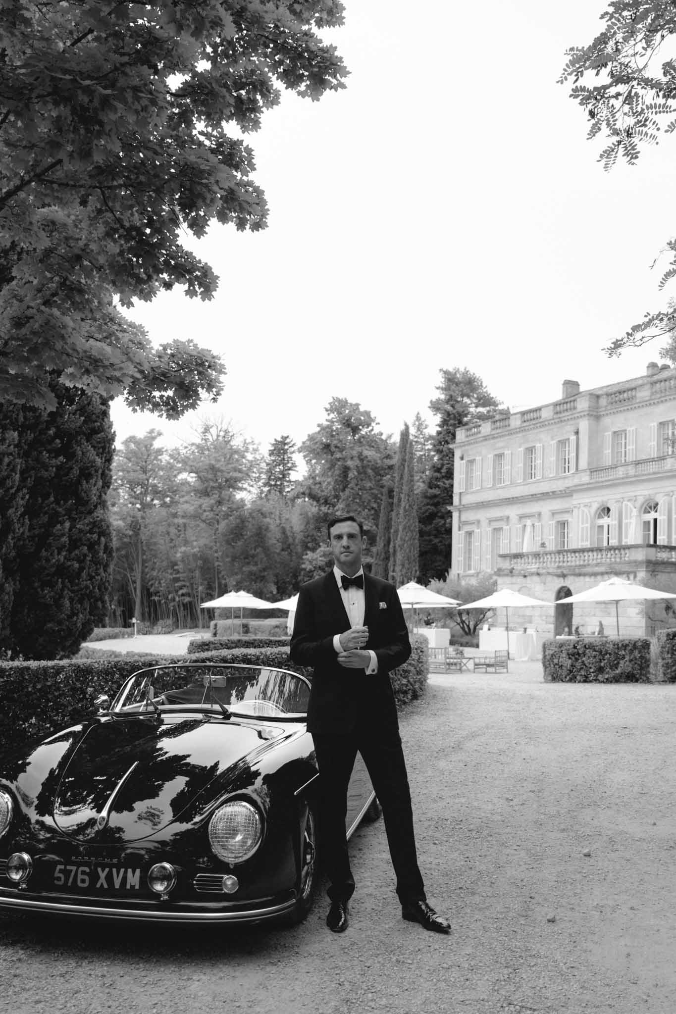 Black-and-white portrait of groom in tuxedo beside vintage convertible in neoclassical estate courtyard