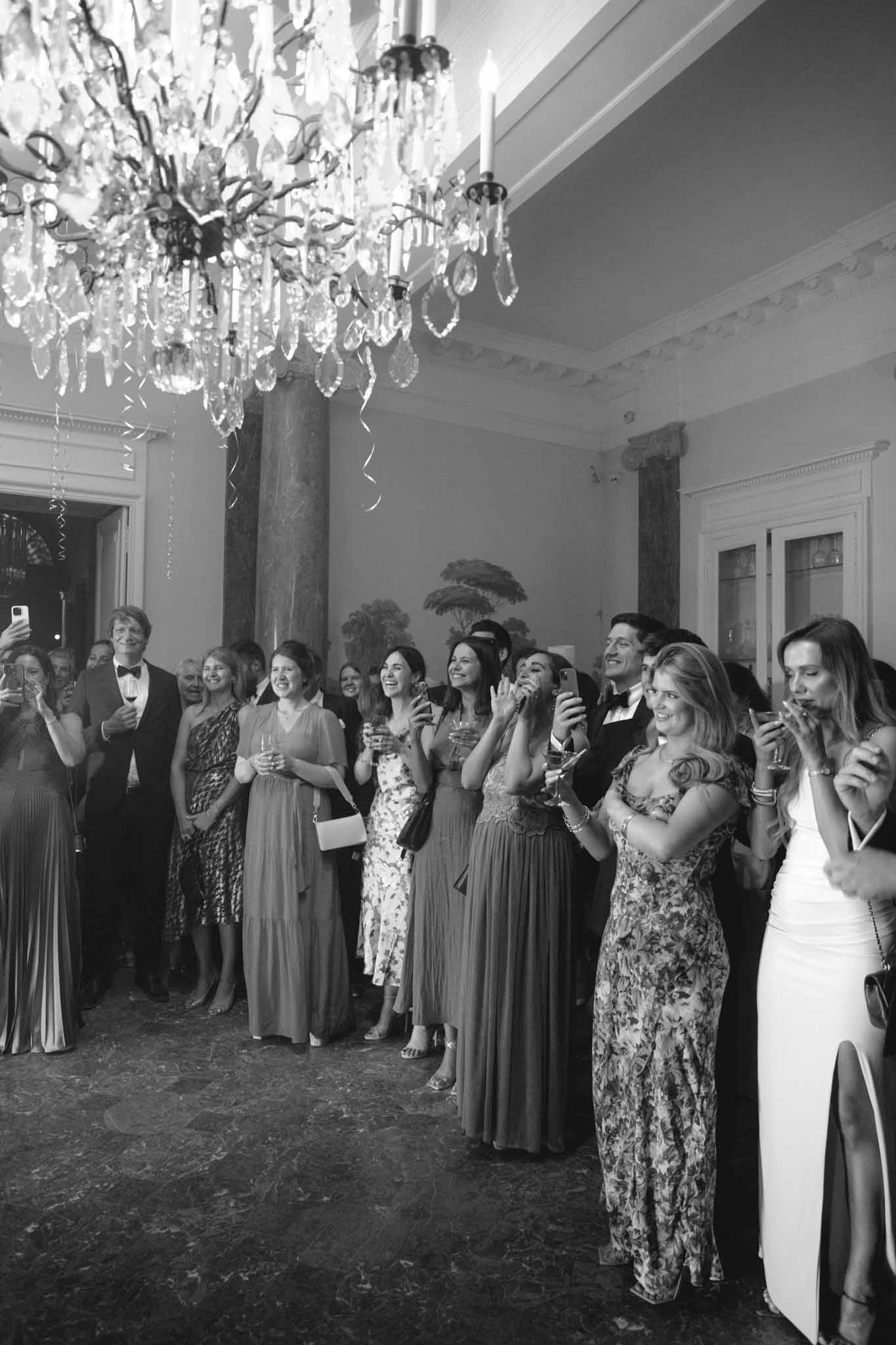 Black-and-white guests in ballroom beneath crystal chandelier watching toast or first dance