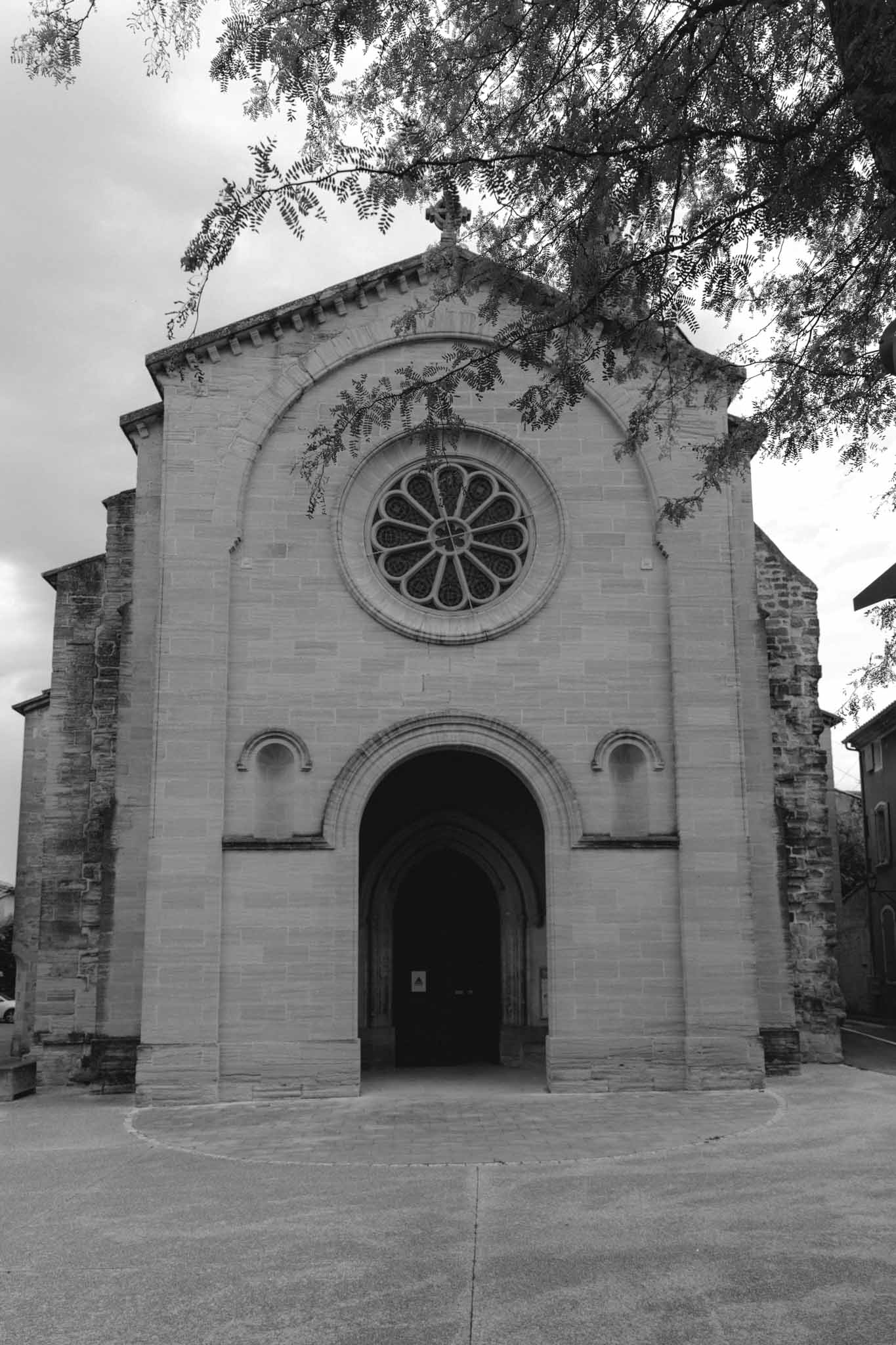 Black-and-white exterior of small stone chapel with arched wooden door and rose window framed by tree branches