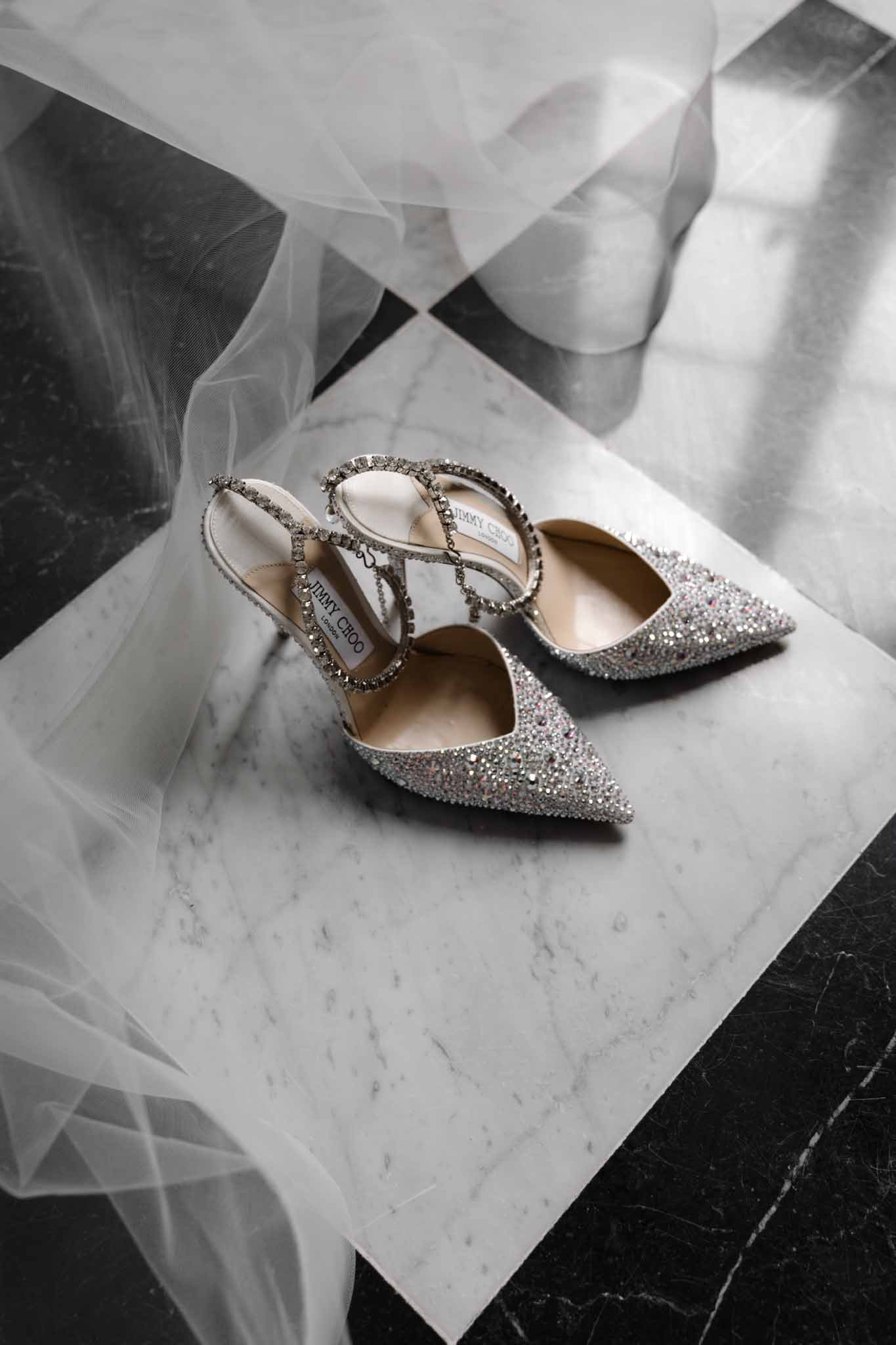 Crystal-embellished pointed-toe bridal heels on white marble with flowing tulle fabric