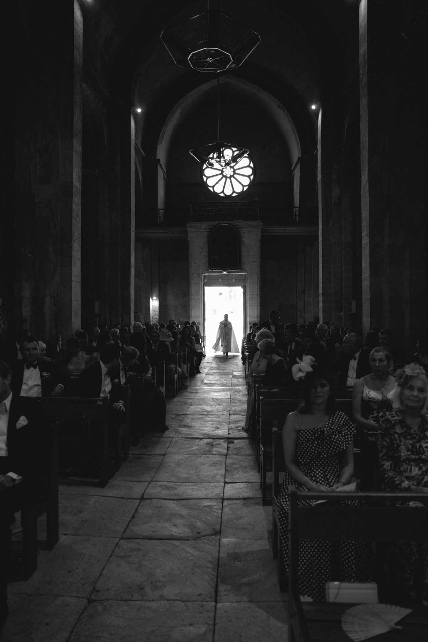 Bride silhouetted in chapel doorway with guests seated in pews under barrel-vaulted ceiling and rose window