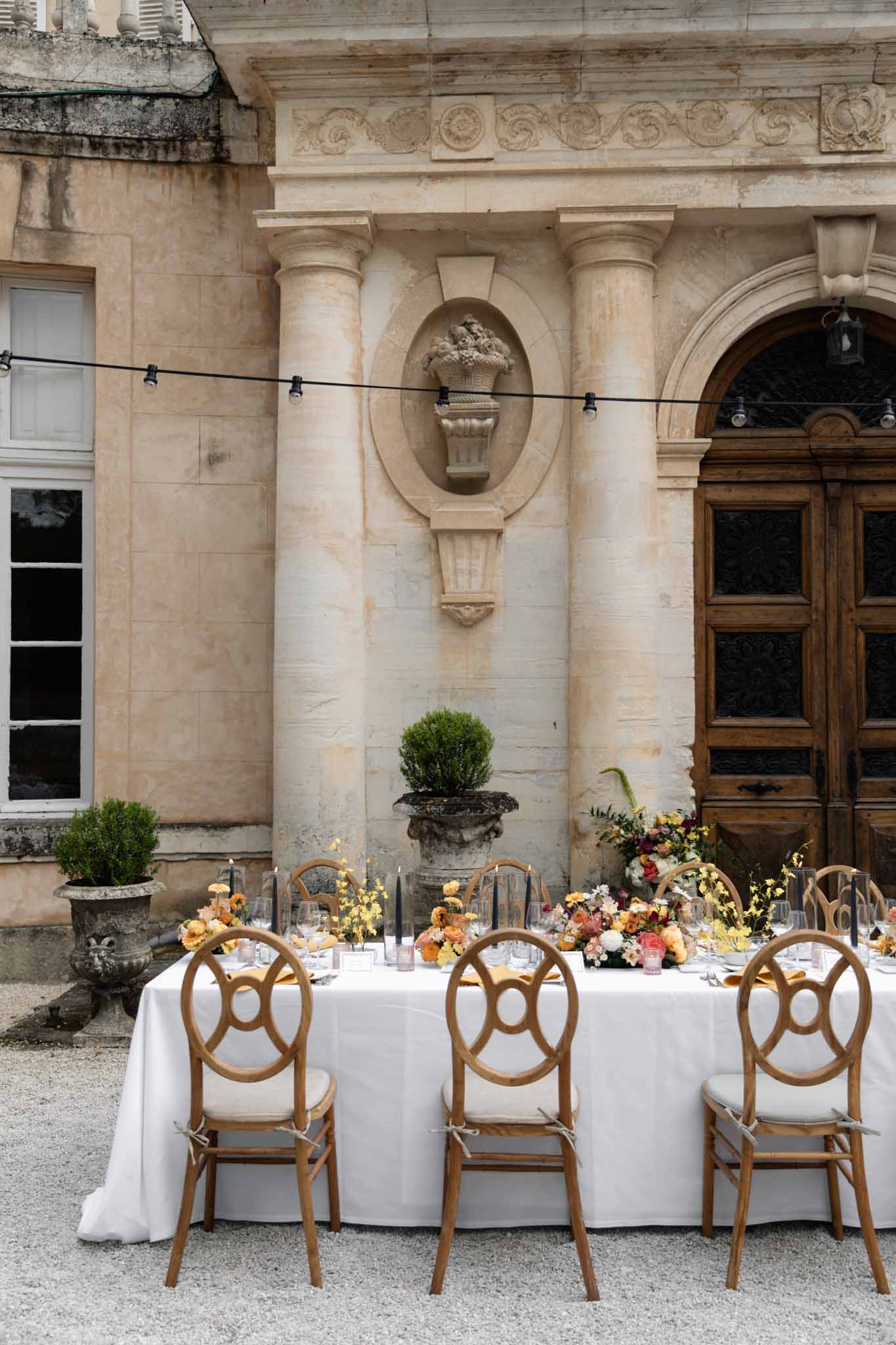 Courtyard reception table with yellow orange and burgundy florals navy taper candles ring-back chairs before chateau facade
