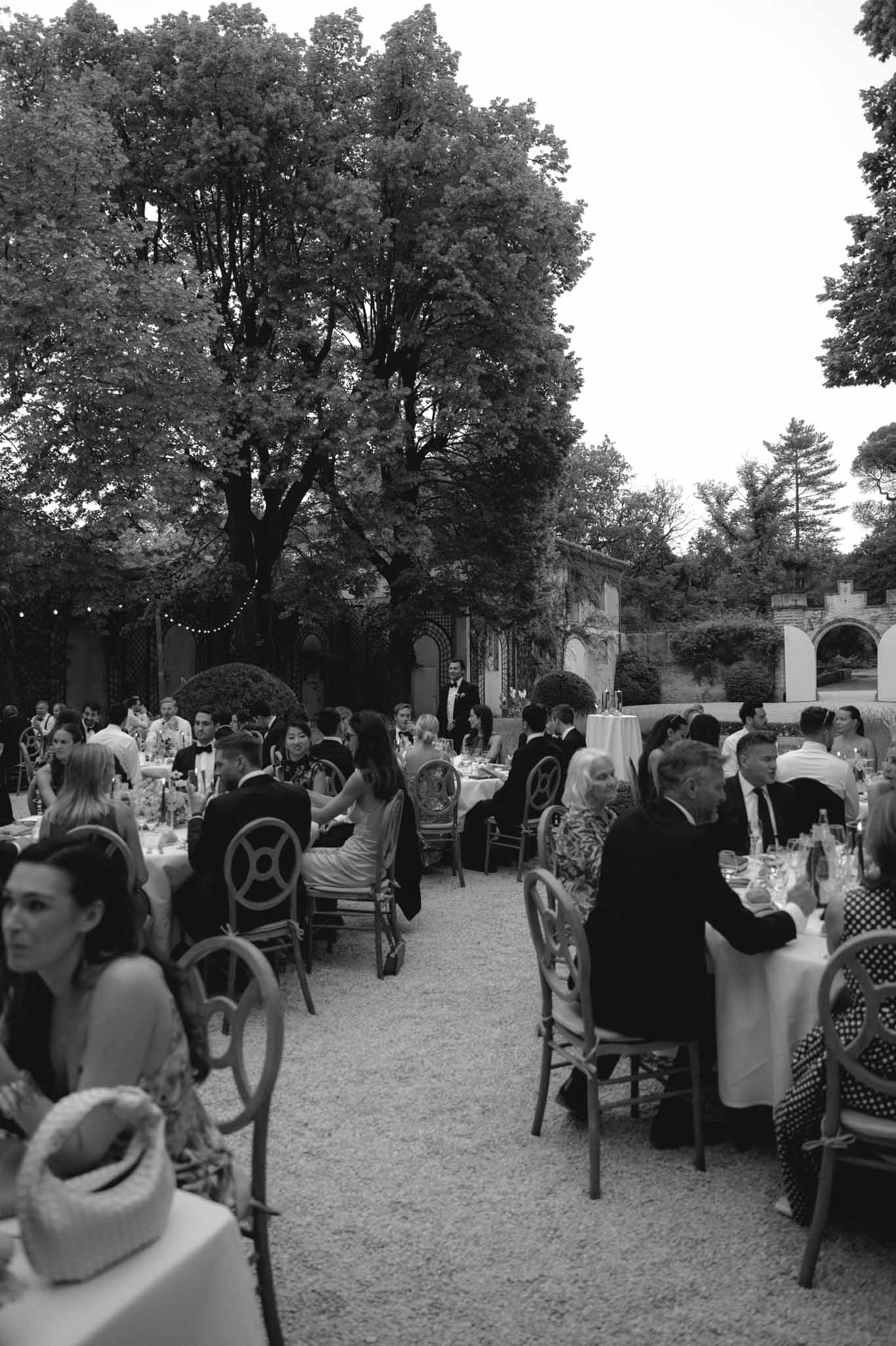 Black-and-white wide shot of 40-50 guests at round tables in stone courtyard with bistro lights and mature trees