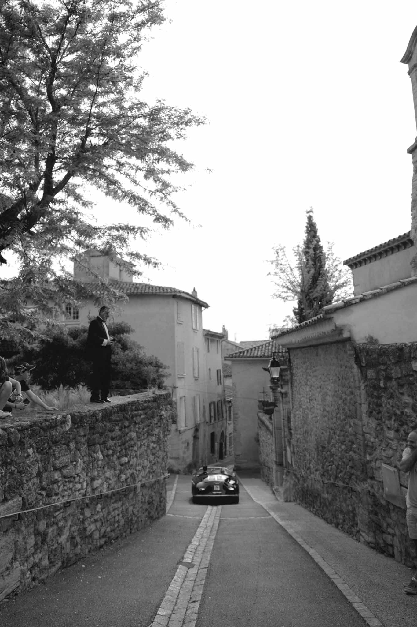 Black and white vintage car driving up cobblestone Provencal village street with stone wall onlookers