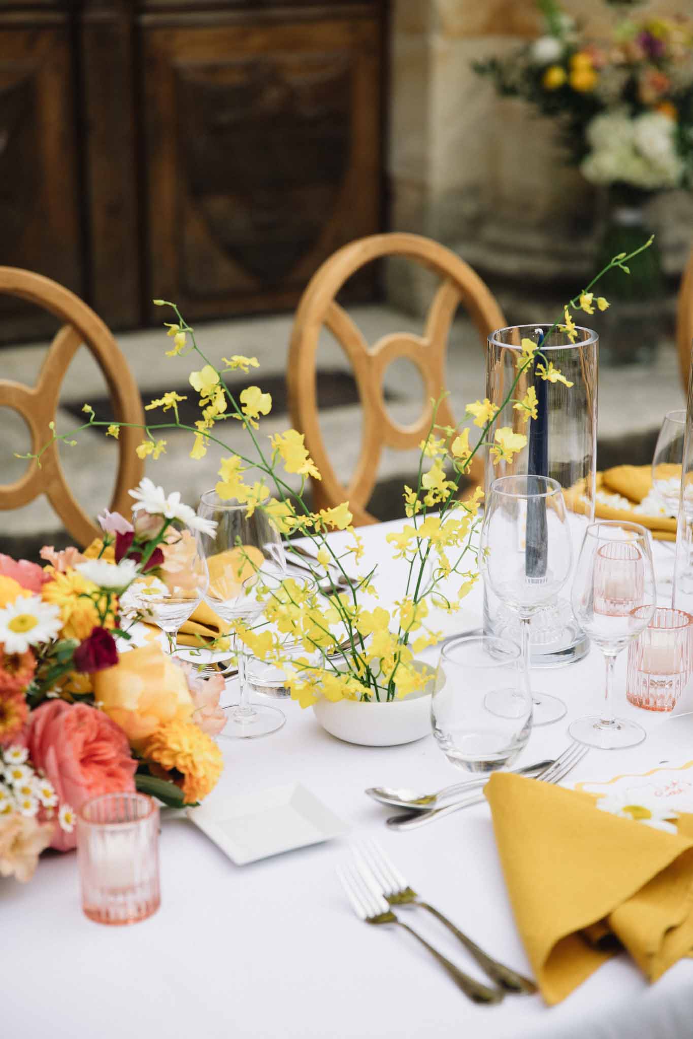 Reception table setting with yellow billy button centerpieces, coral roses, and white linens on a long dining table