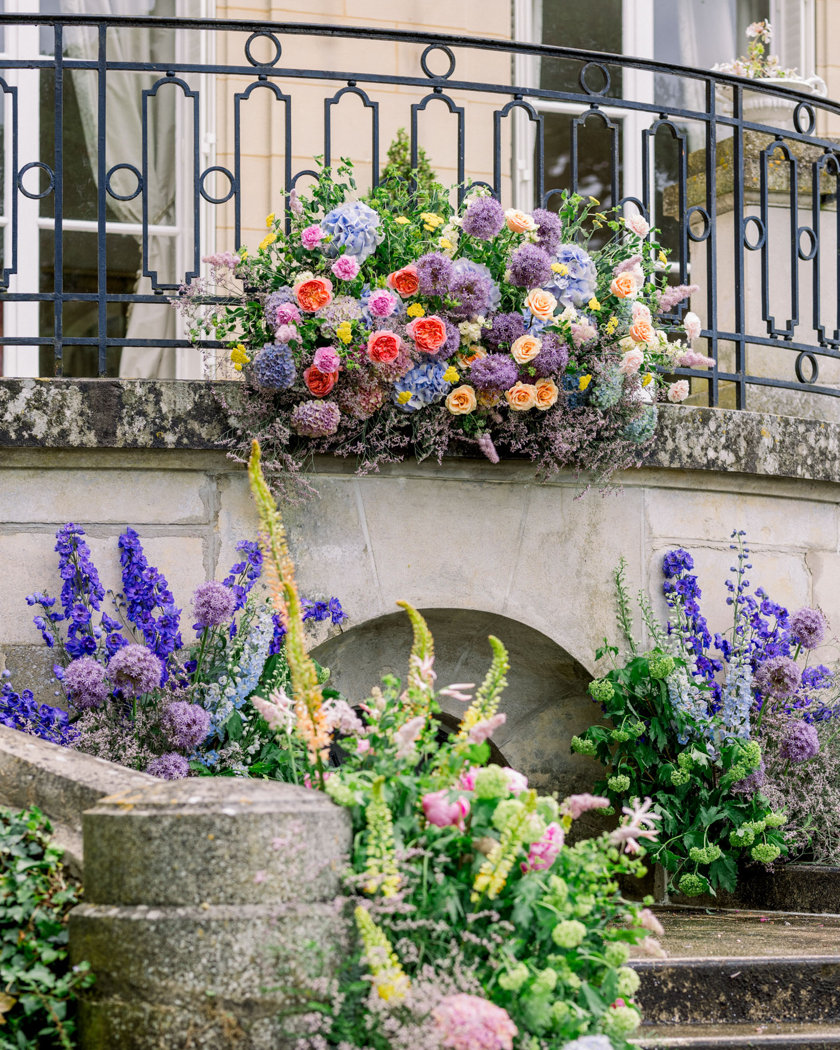 Garden-style floral installation on chateau stone staircase with coral roses, blue hydrangeas, and purple alliums