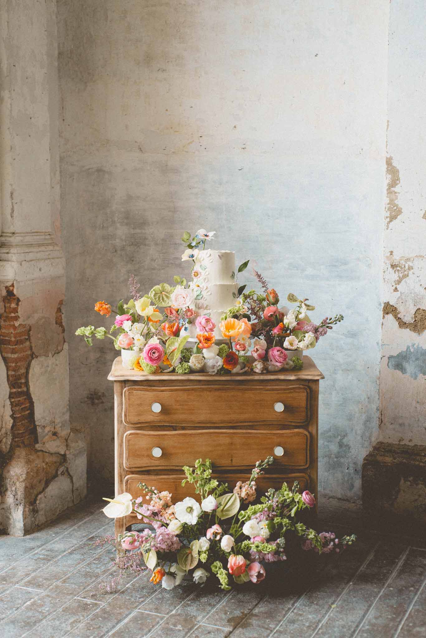 A wedding cake display styled on a vintage wooden chest of drawers with white ceramic knob handles, photographed as a detail shot indoors. The two-tier ivory cake features textured buttercream frosting with small hand-painted or sugar flower details and is topped with blue and white anemone blooms. The cake is surrounded by a loose, garden-style floral arrangement covering the top of the dresser and cascading down to the floor, incorporating orange poppies, hot pink and blush ranunculus, coral roses, white anemones, anthurium leaves, astilbe, green amaranthus, and mixed foliage in a vivid, painterly color palette. A secondary floral cluster is arranged on the floor in front of the dresser with similar blooms including white anemones, pink tulips, lavender stock, and trailing greenery. The backdrop is a heavily distressed plaster wall with patches of pale blue paint and exposed brick, giving the setting a rustic, aged-interior atmosphere.