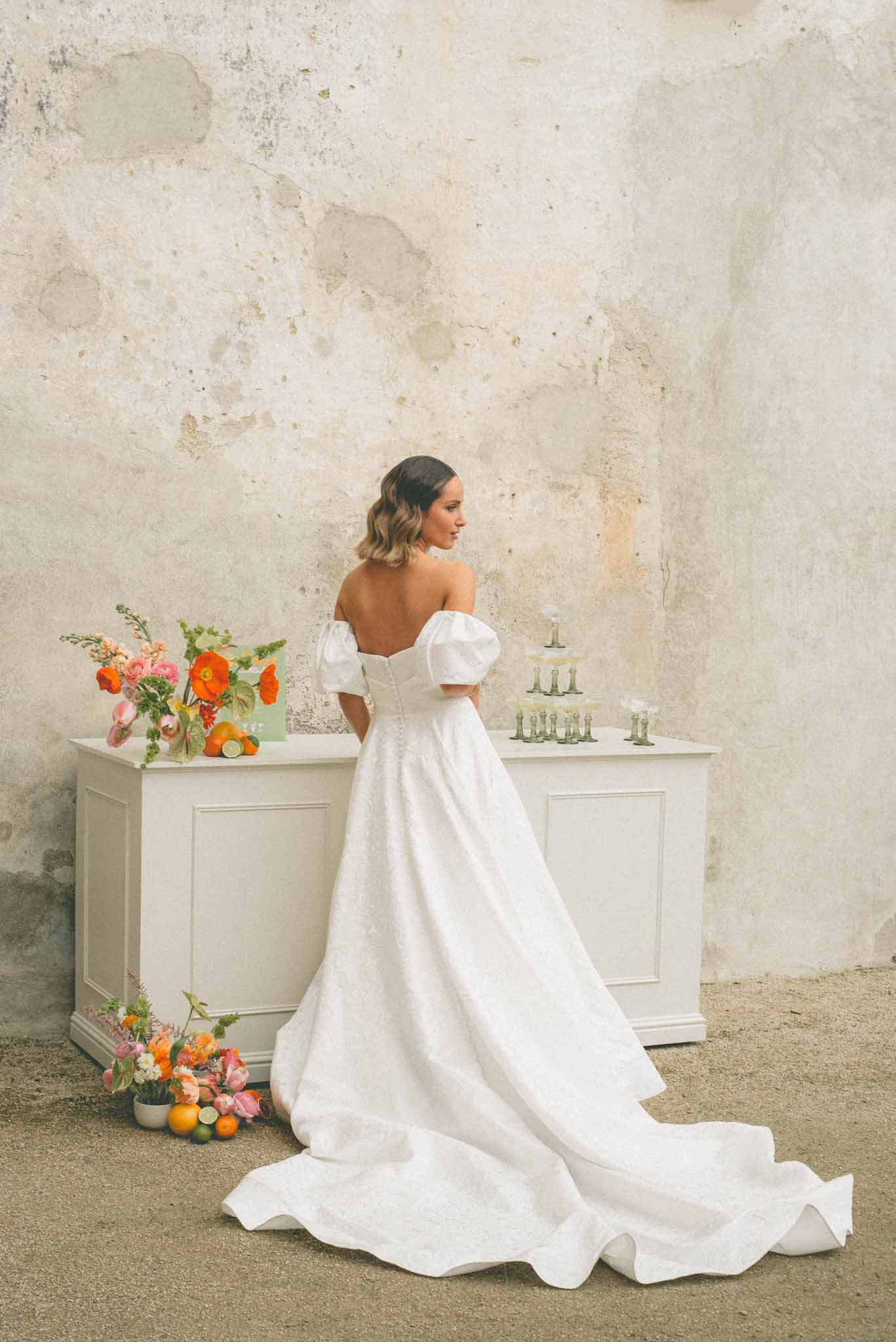 Bride in puff-sleeve corseted gown from behind beside orange poppy and pink ranunculus bar arrangement