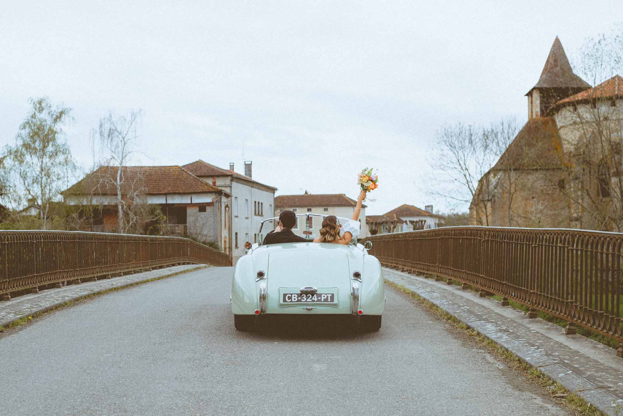 Couple driving away in mint-green vintage convertible bride raising colourful bouquet crossing village bridge