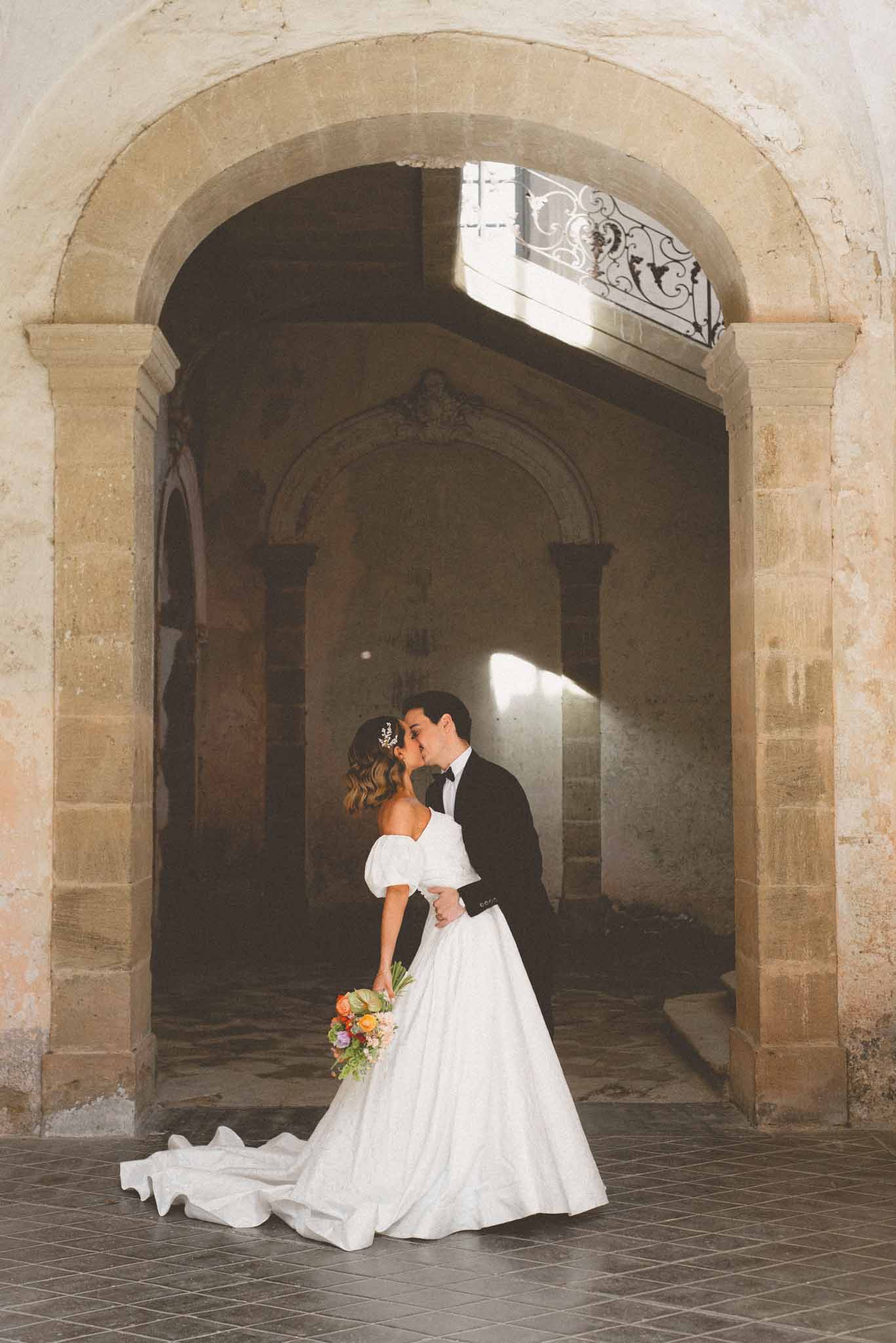Bride and groom kissing beneath stone archway inside historic chateau with wrought-iron staircase